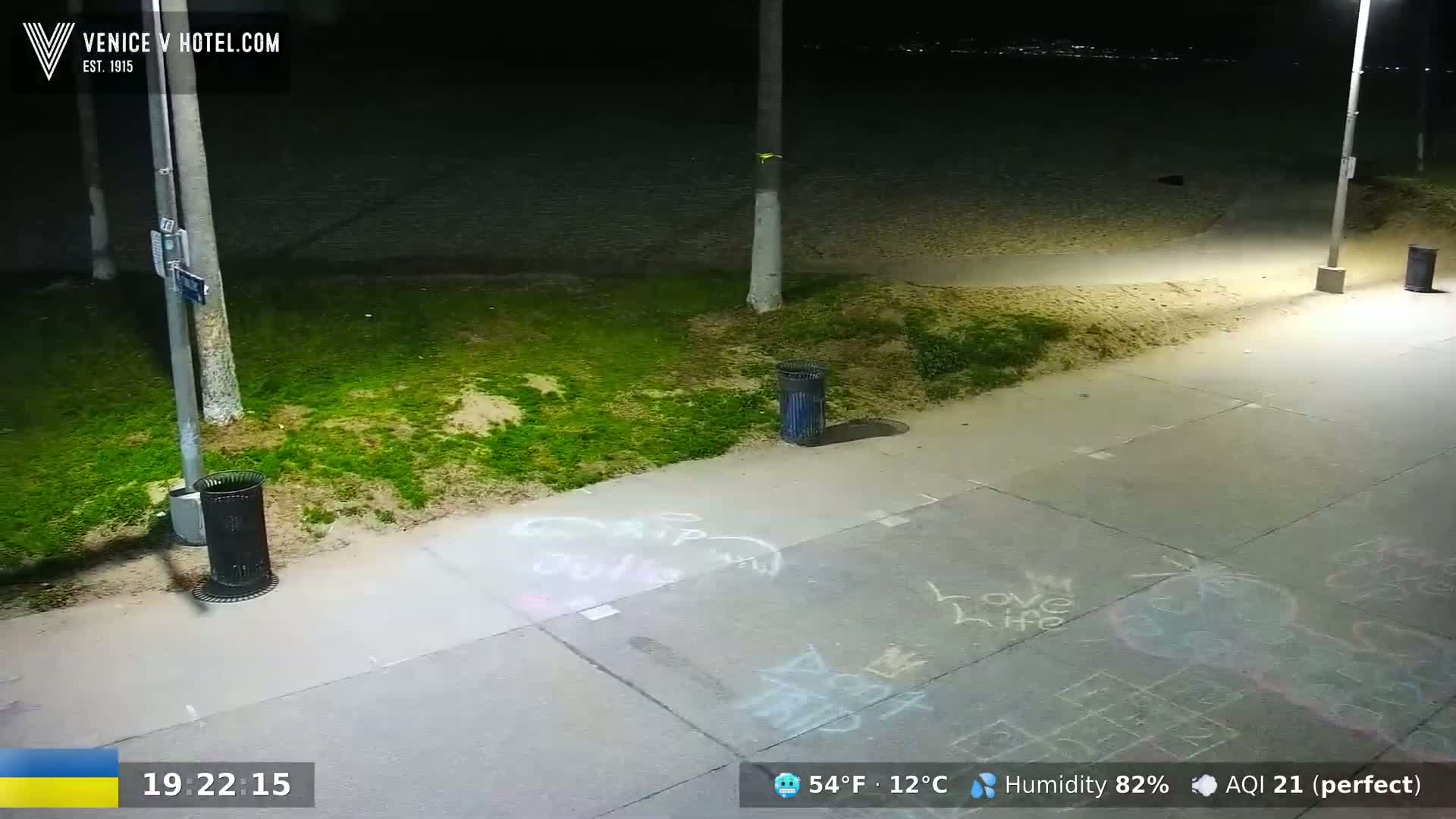 A nighttime beachside scene shows a paved walkway with chalk drawings, trash cans, and grassy areas under partly cloudy conditions.