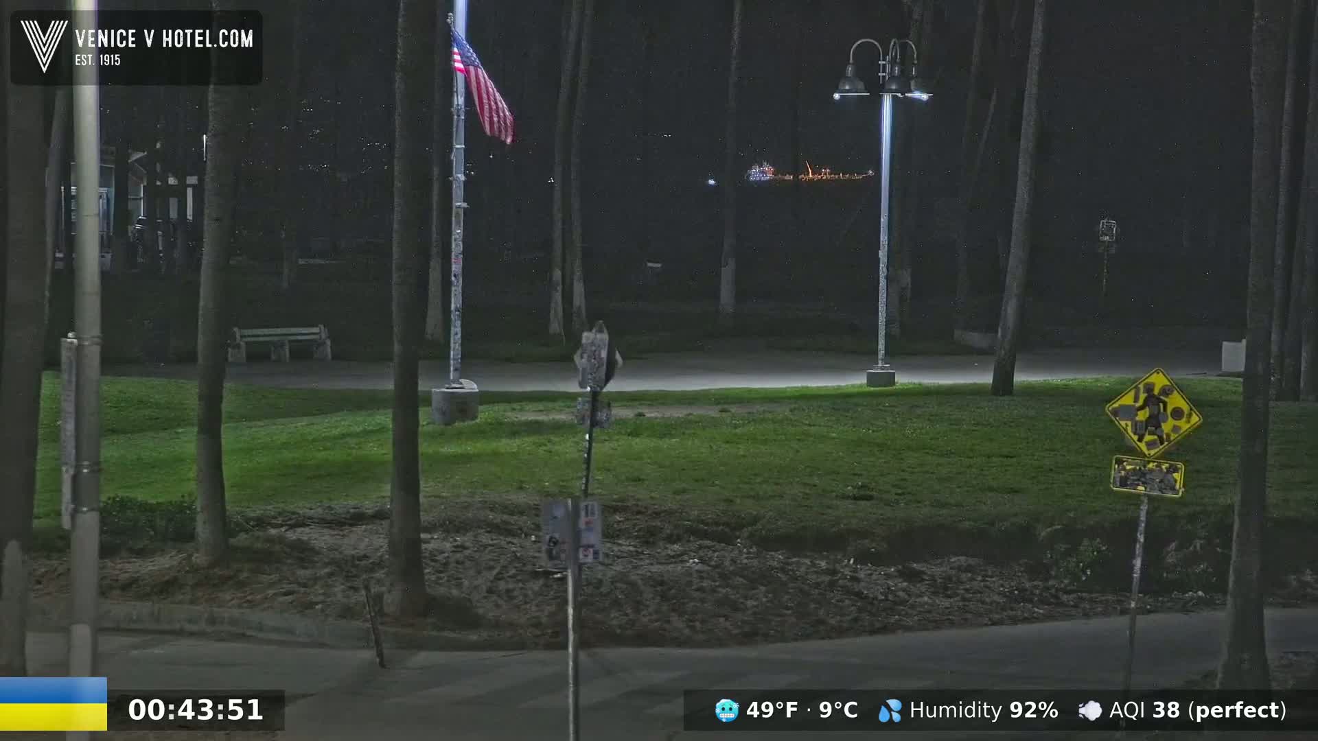 A nighttime view of a park with a flagpole displaying an American flag, streetlights, and a grassy area under a clear sky.