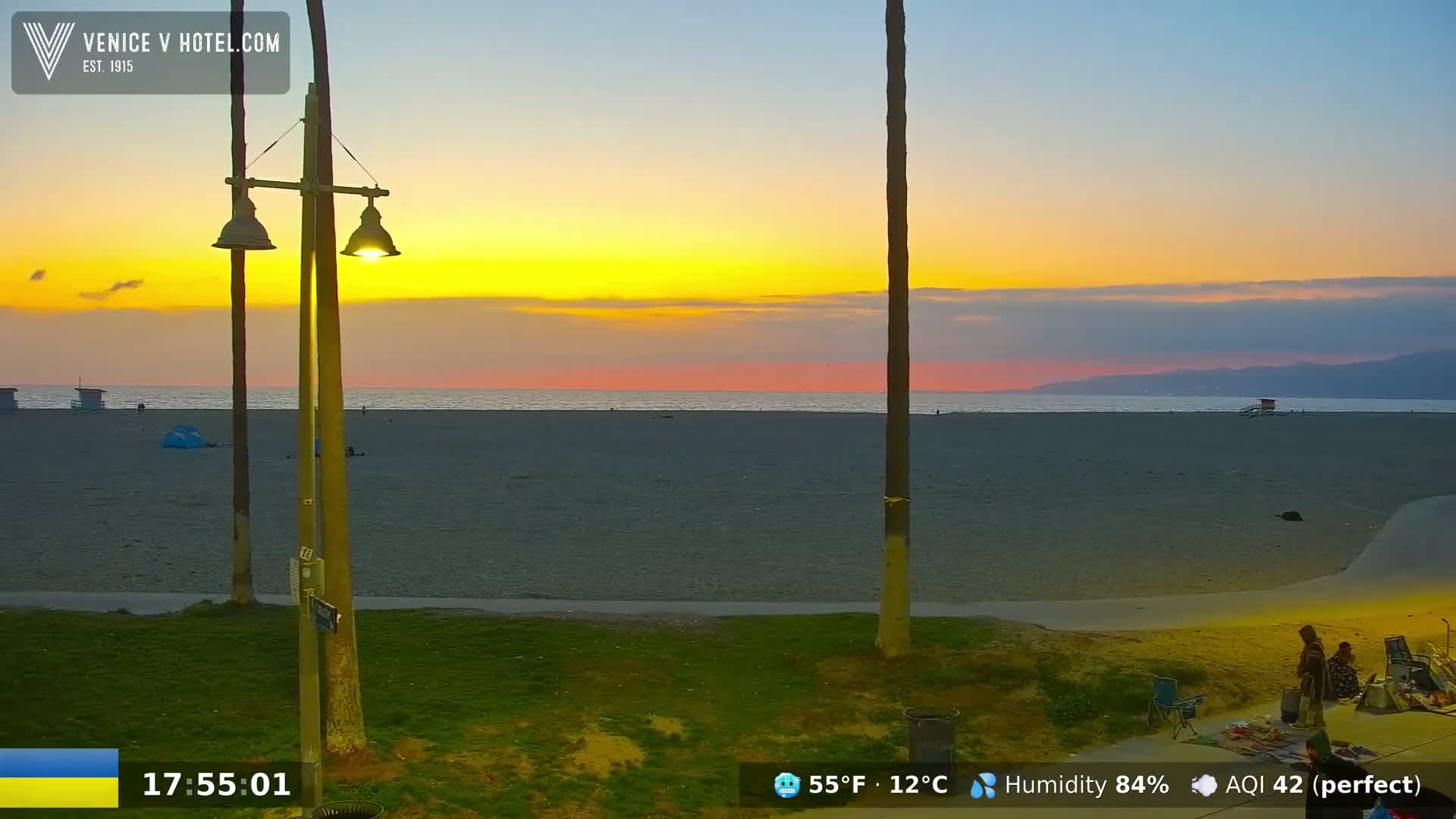 A beach scene at sunset shows a mostly empty sandy beach with a few people near a lamppost and palm trees, under a partly cloudy sky.