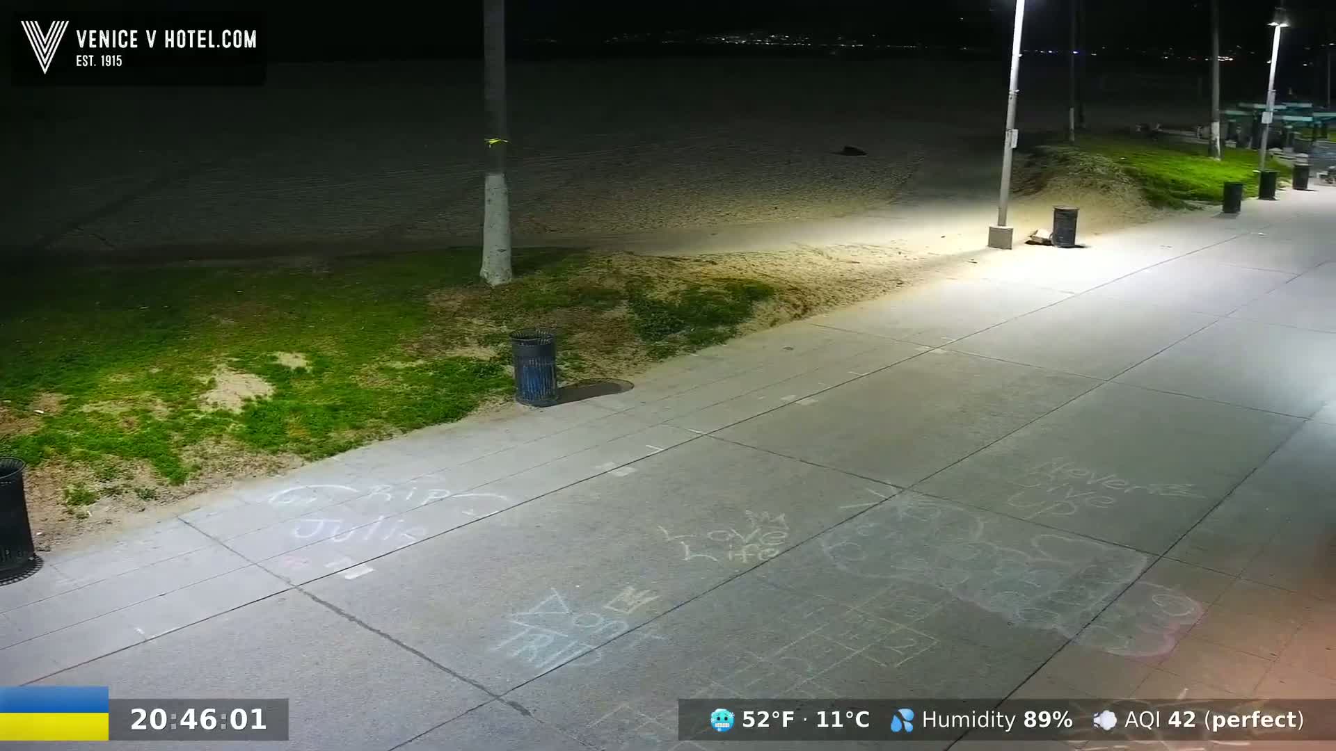 A nighttime view of a paved walkway next to a sandy beach, illuminated by streetlights, with chalk writings on the ground.