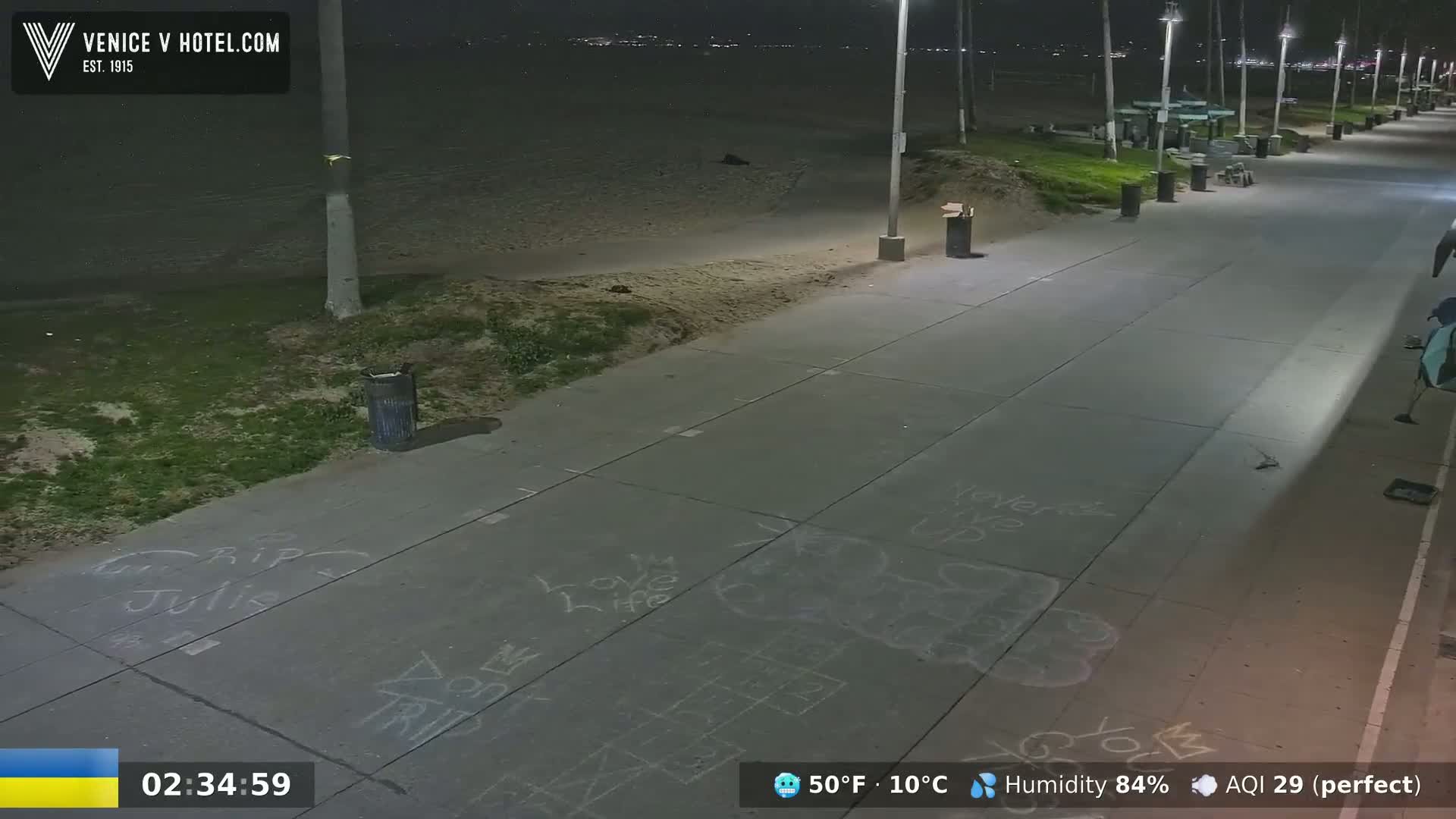 A nighttime view of a beach boardwalk shows chalk drawings on the pavement, with a sandy beach and ocean visible in the background under a clear sky.