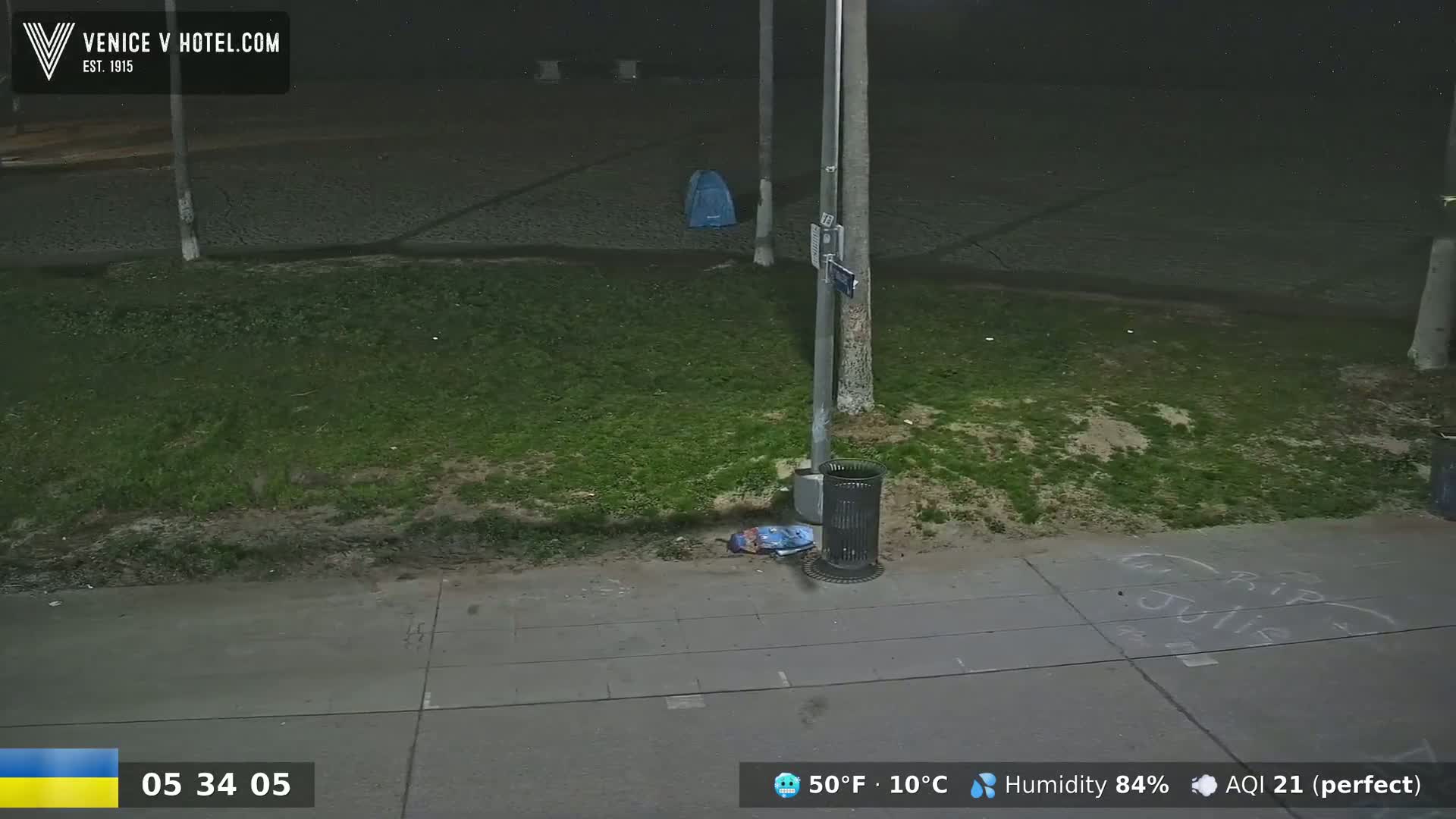 A nighttime, outdoor scene shows a sidewalk, grassy area, a trash can, and a blue object on the ground near a streetlamp under a dark sky.