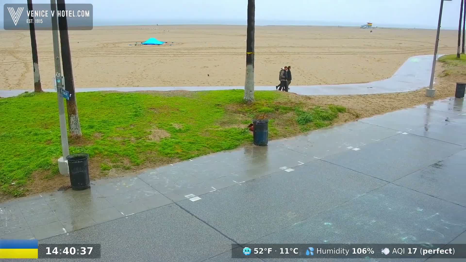 A wet beach scene shows a sandy beach with a few people walking on a nearby paved walkway, under an overcast sky.