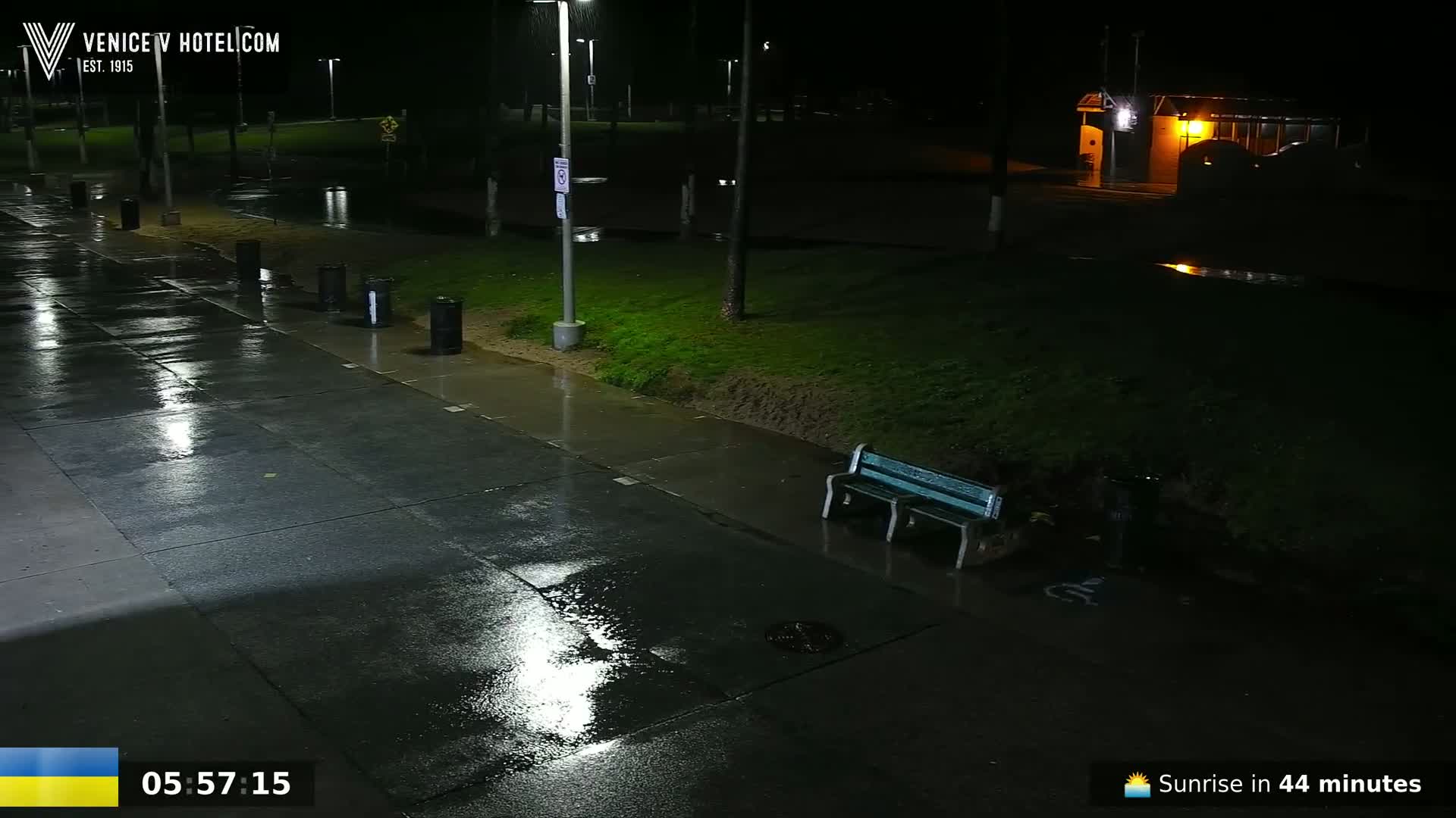 A wet, nighttime scene shows a rain-slicked walkway next to a grassy area with a bench under a lamppost.