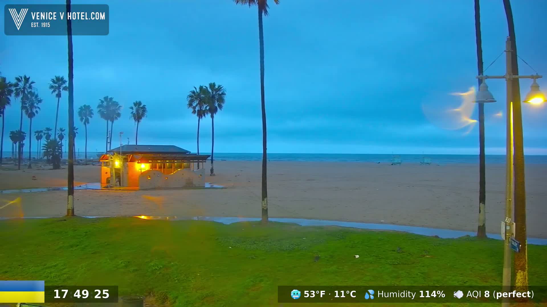 A beach scene at dusk shows a small illuminated building on a sandy beach with palm trees and a grassy area, under an overcast sky.