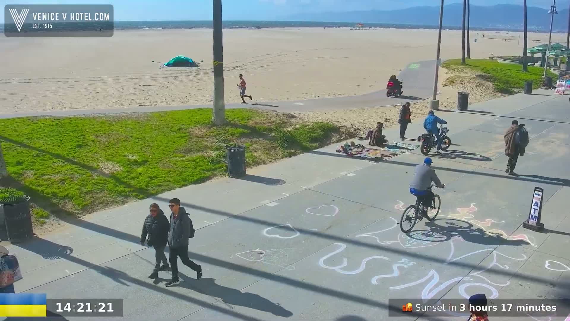 A sunny beach scene shows people walking and biking along a paved path next to a sandy beach, with chalk drawings on the path.