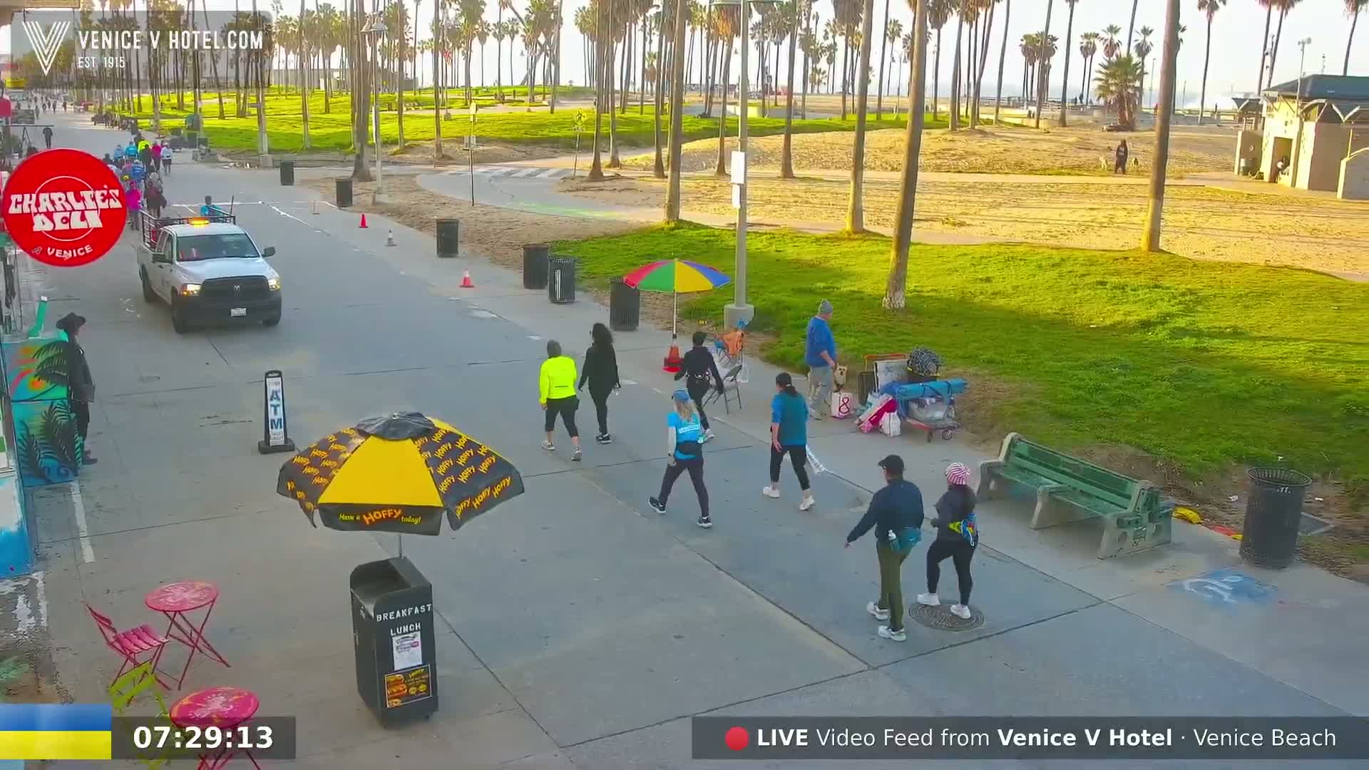 A sunny morning scene on a paved beachfront walkway shows several pedestrians walking, a food vendor with an umbrella, a truck parked on the side of the road, and palm trees lining the sandy beach in the background.