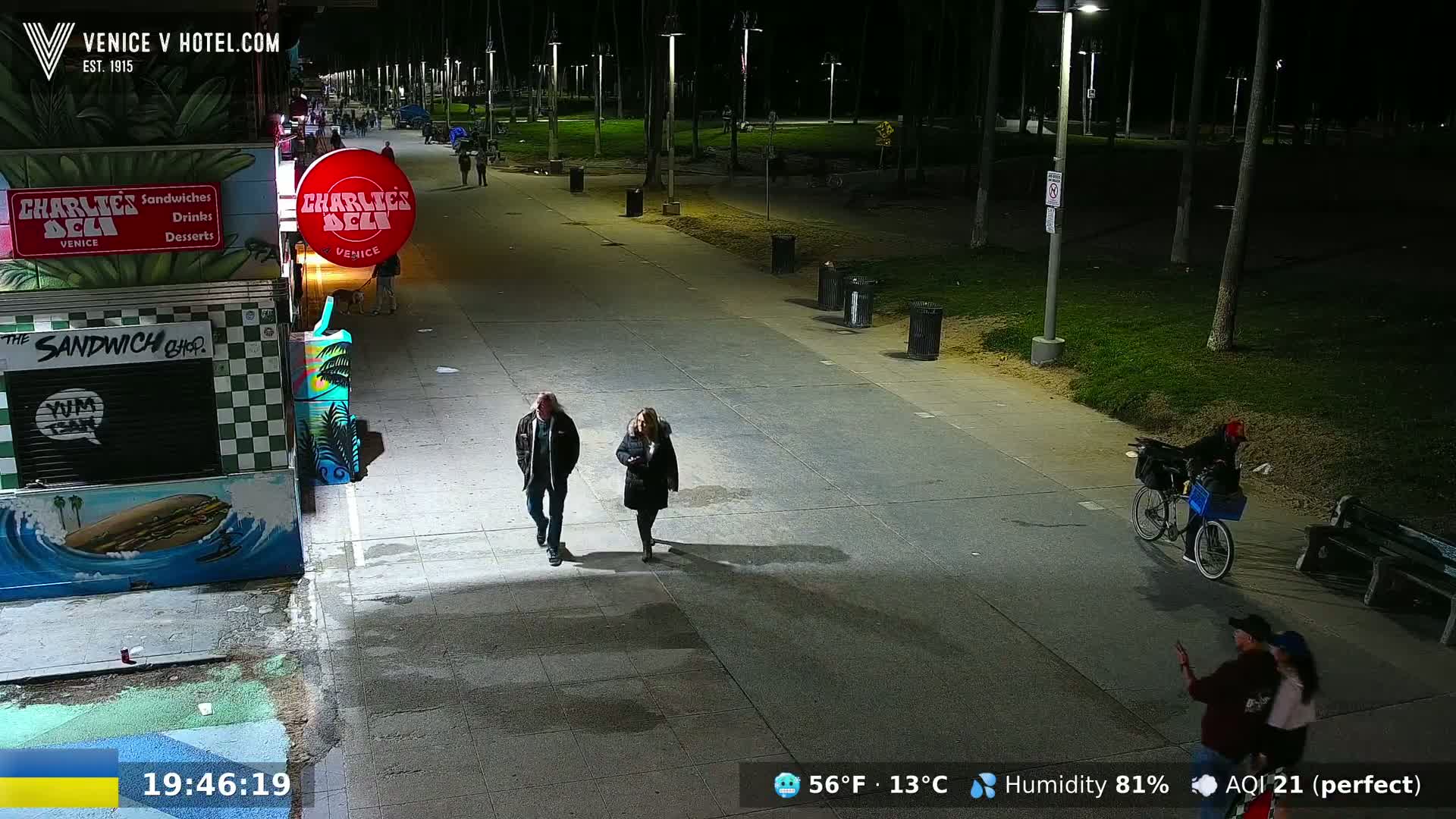 A nighttime, outdoor scene shows a paved walkway with people walking, a person riding a bicycle, and several trash cans, all under well-lit streetlights.