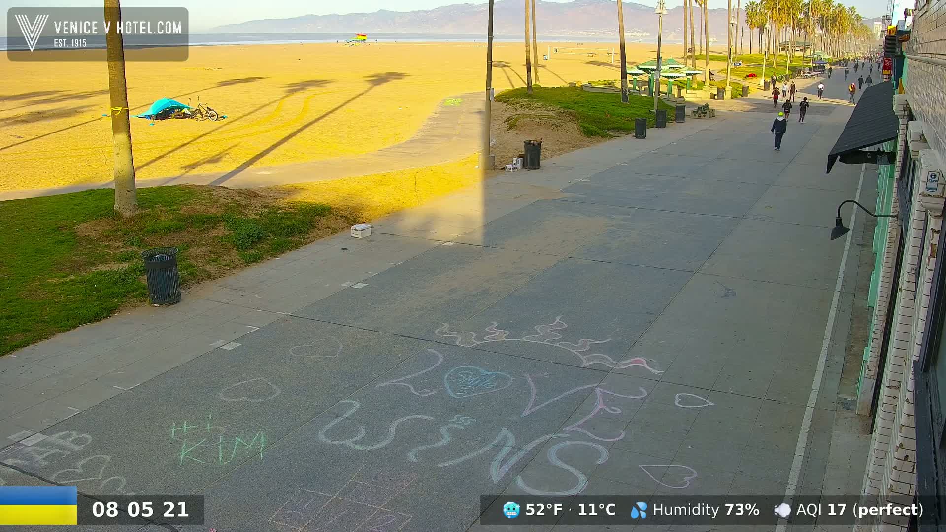 A sunny beach boardwalk scene shows people walking, a sandy beach with palm trees casting shadows, and chalk drawings on the pavement.