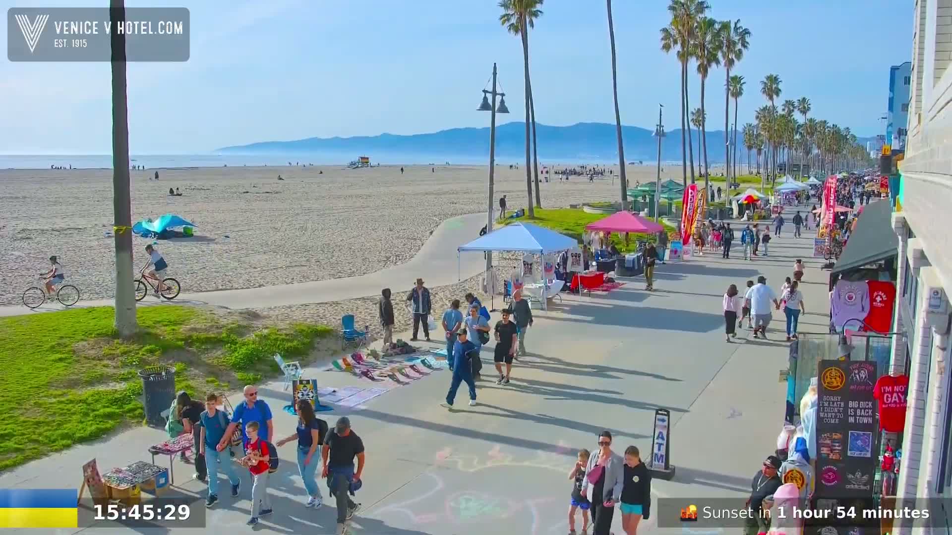 A sunny beach boardwalk scene shows people walking, vendors selling goods under pop-up tents, and cyclists riding along a paved path next to a sandy beach with calm ocean waves.