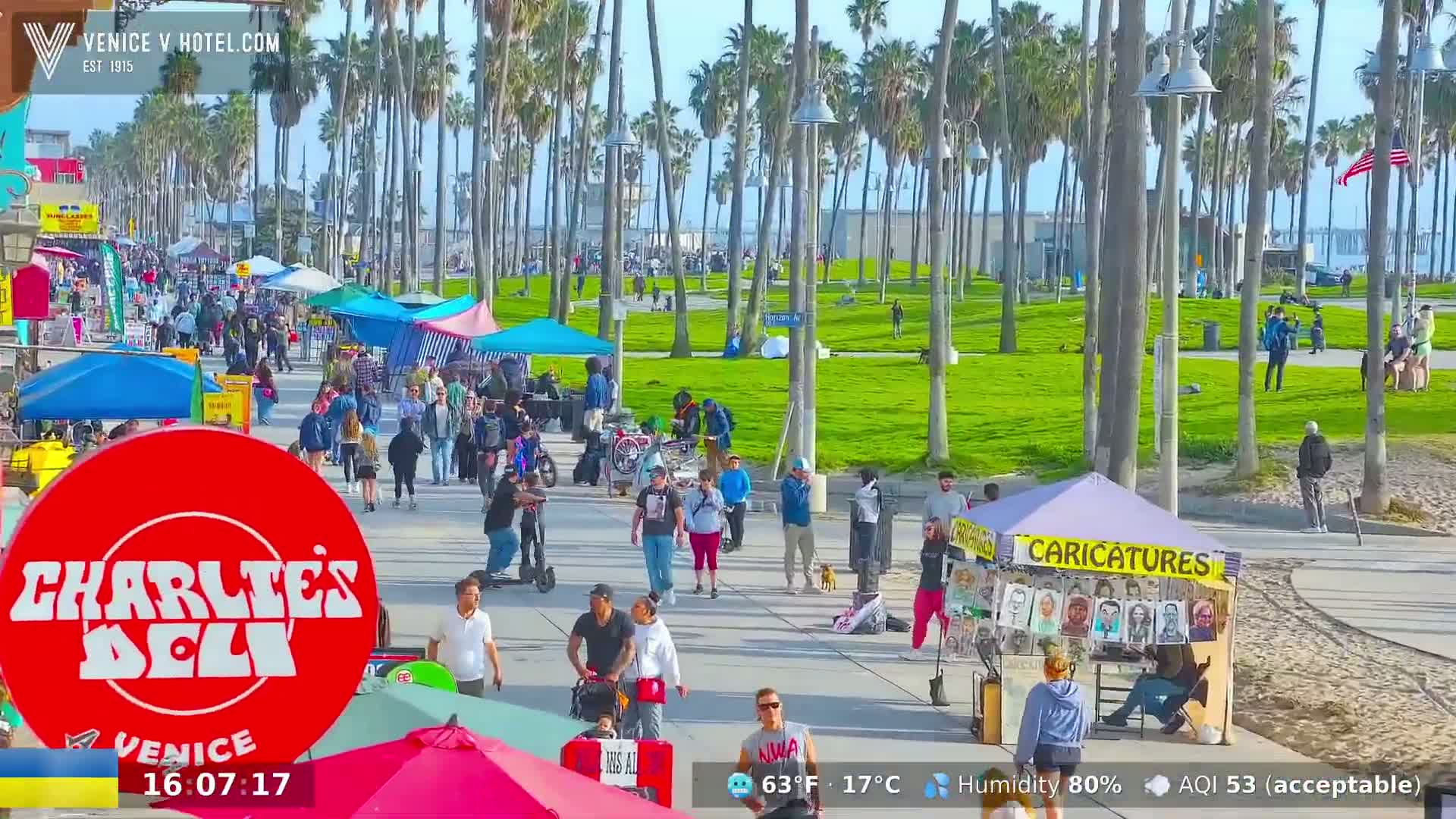 A sunny boardwalk scene with many people walking past various vendors and palm trees, under a partly cloudy sky.