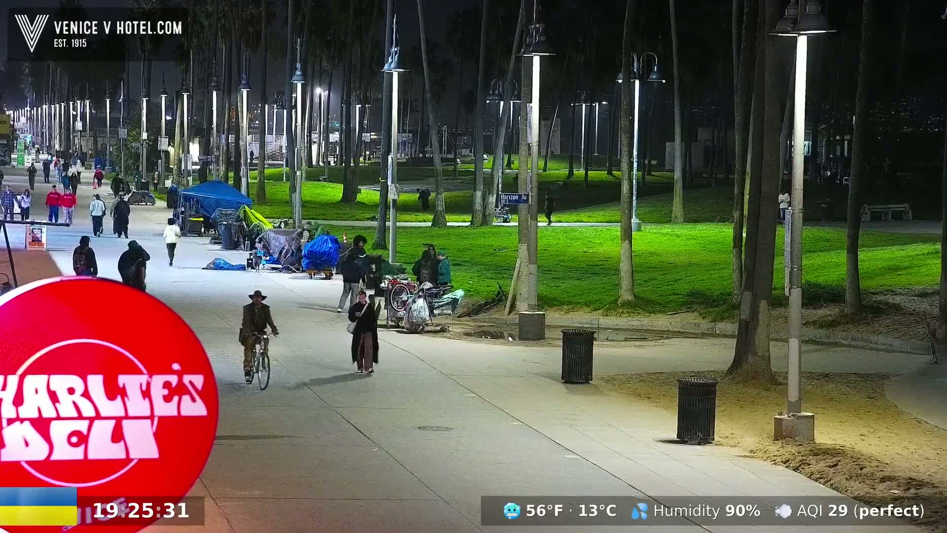 A nighttime scene shows a paved walkway lined with palm trees and streetlights, with people walking and biking, and some tents and belongings suggesting homelessness, under a humid sky with good air quality.