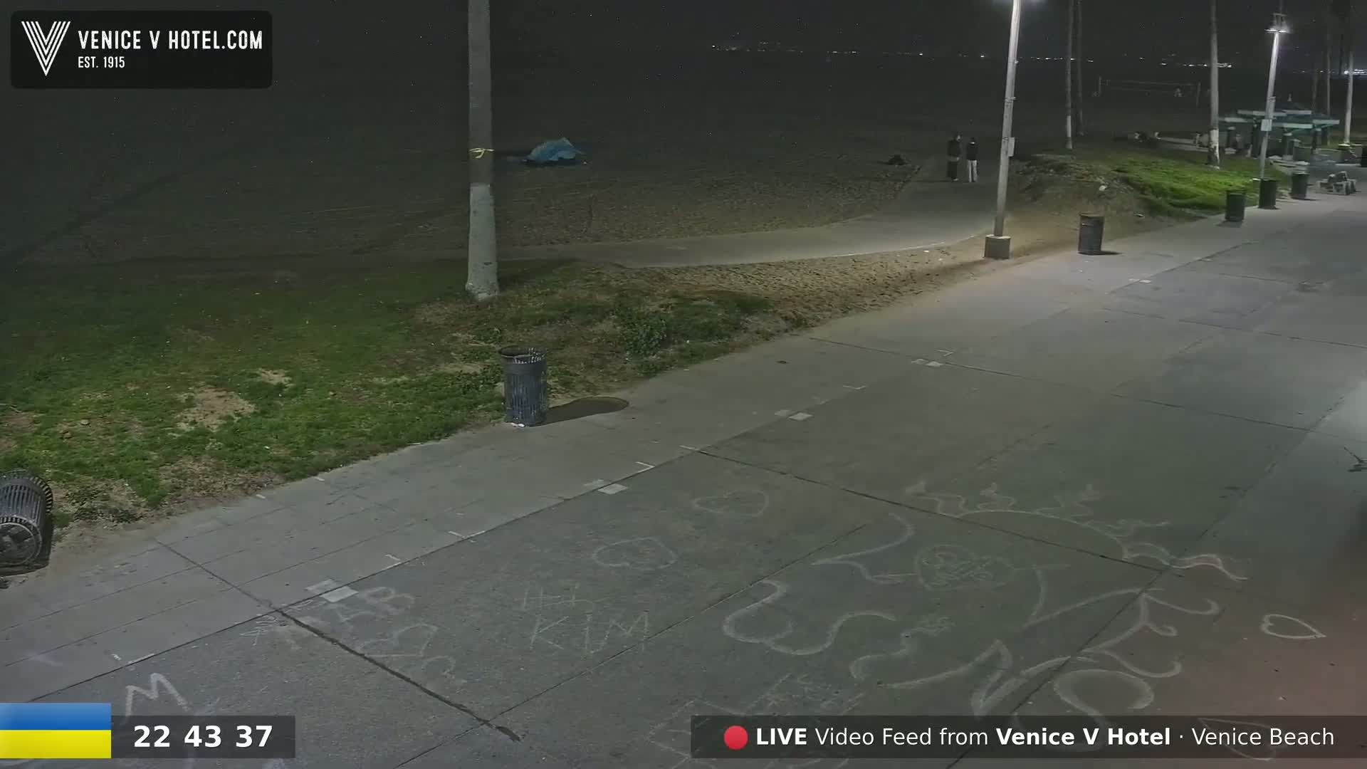 A nighttime view of a paved walkway next to a beach, illuminated by streetlights, showing some drawings on the ground and a few people in the distance.