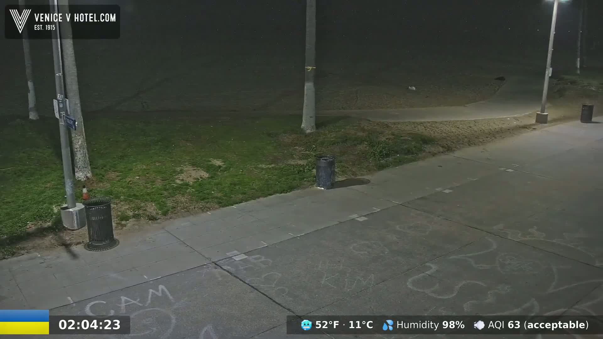 A nighttime scene shows a sidewalk with graffiti, a grassy area, trash cans, and a paved path leading to a sandy beach under hazy conditions.