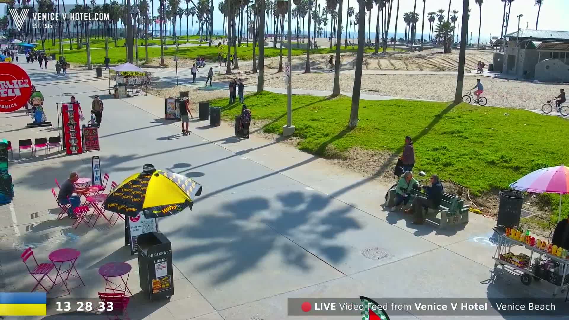 A sunny beach boardwalk scene shows people sitting at tables under umbrellas, walking along a paved path, and riding bikes near a grassy area with palm trees, next to a sandy beach.