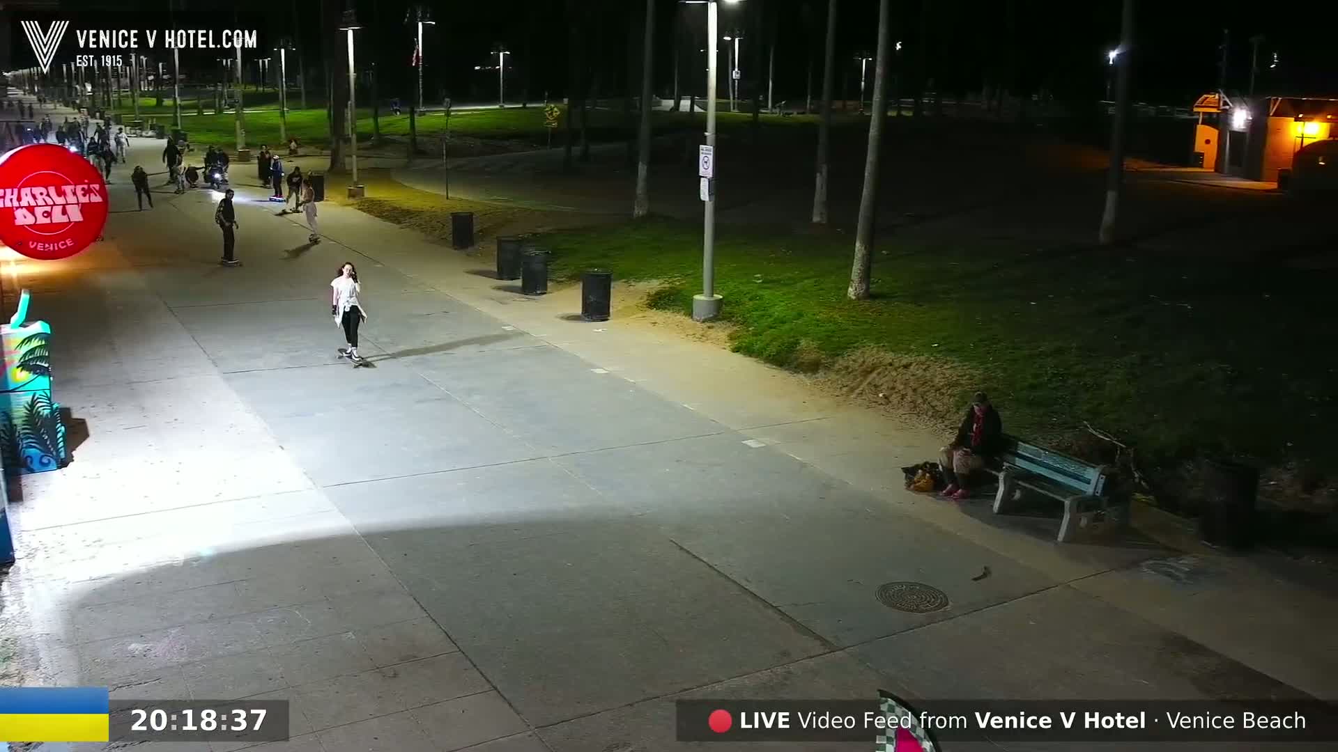 A nighttime scene shows several people skateboarding and one person sitting on a bench along a paved walkway next to a grassy area under bright streetlights.