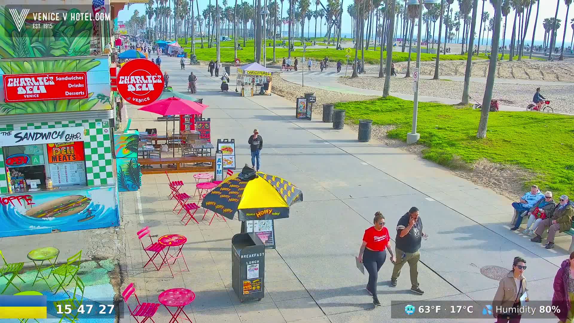 A sunny boardwalk scene shows people walking and sitting near food stands and shops, under a partly cloudy sky.