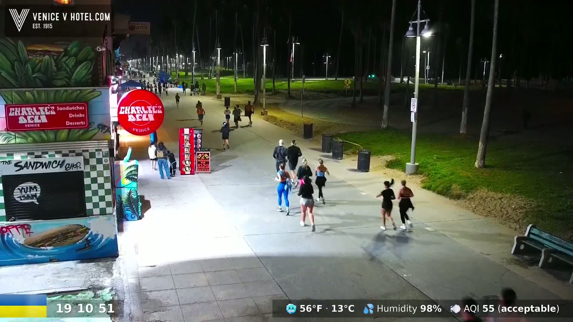 A nighttime scene shows several people jogging along a paved path lined with palm trees, near buildings with outdoor food service.