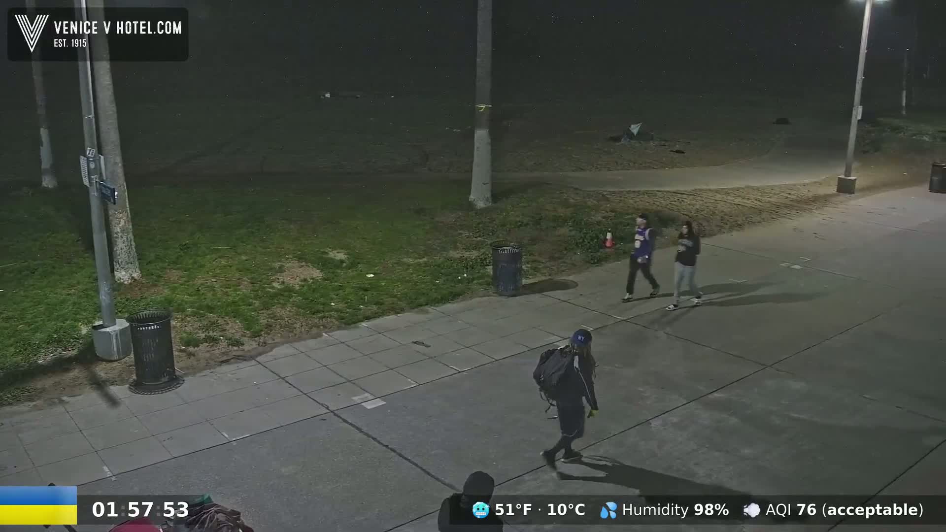 A nighttime, outdoor scene shows three people walking on a sidewalk near a grassy area with trash cans, under a cloudy sky.