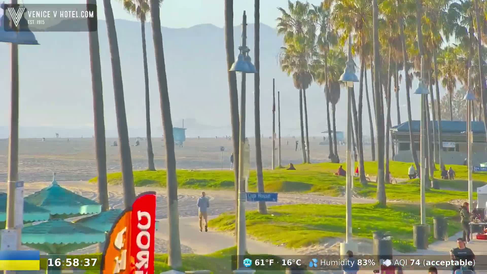 A mostly sunny beach scene shows a wide sandy beach with people on it, palm trees lining a paved walkway, and green grass between the walkway and the beach.