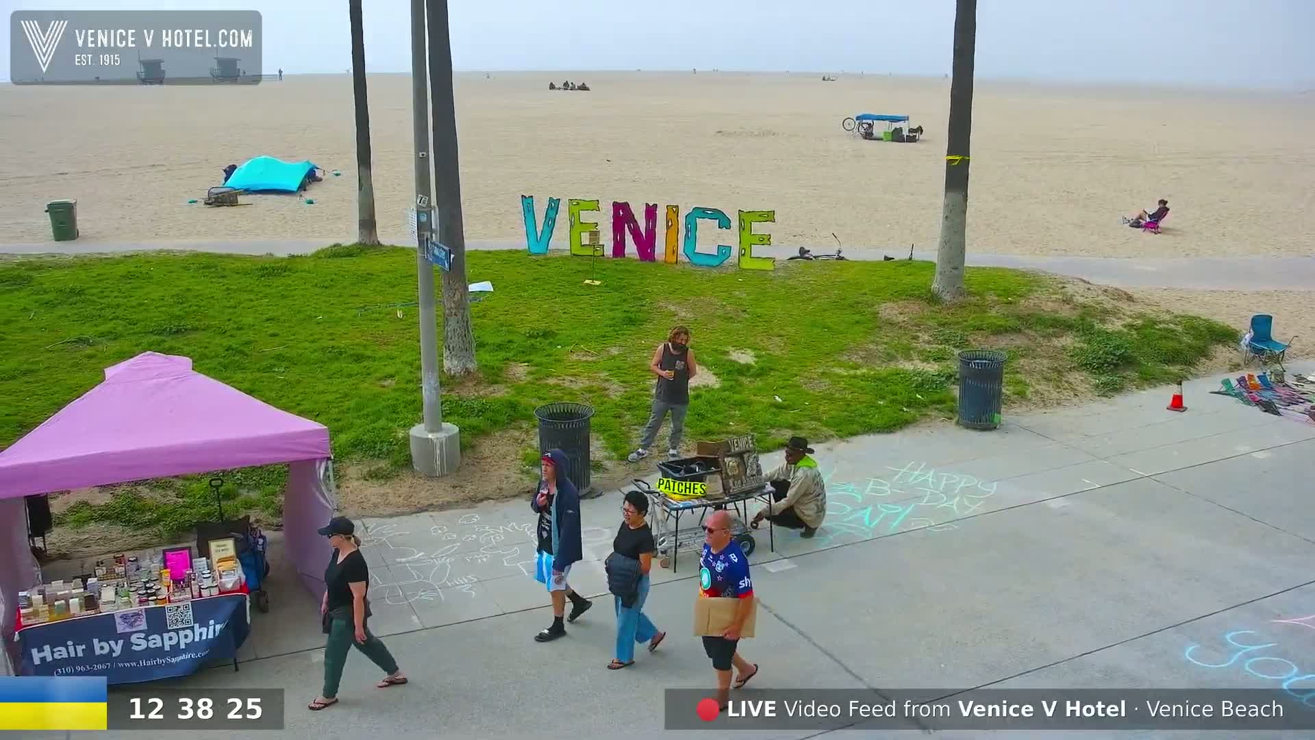 A beach scene on an overcast day shows people milling around near a large "Venice" sign, some vendors' stalls, and chalk drawings on the boardwalk.