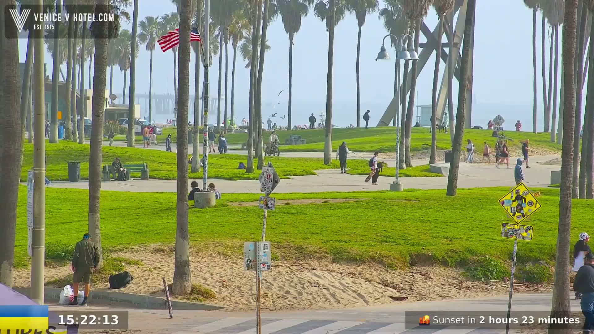A sunny, hazy day at a beachside park shows people walking, sitting, and riding bikes amidst palm trees, with the ocean visible in the background.