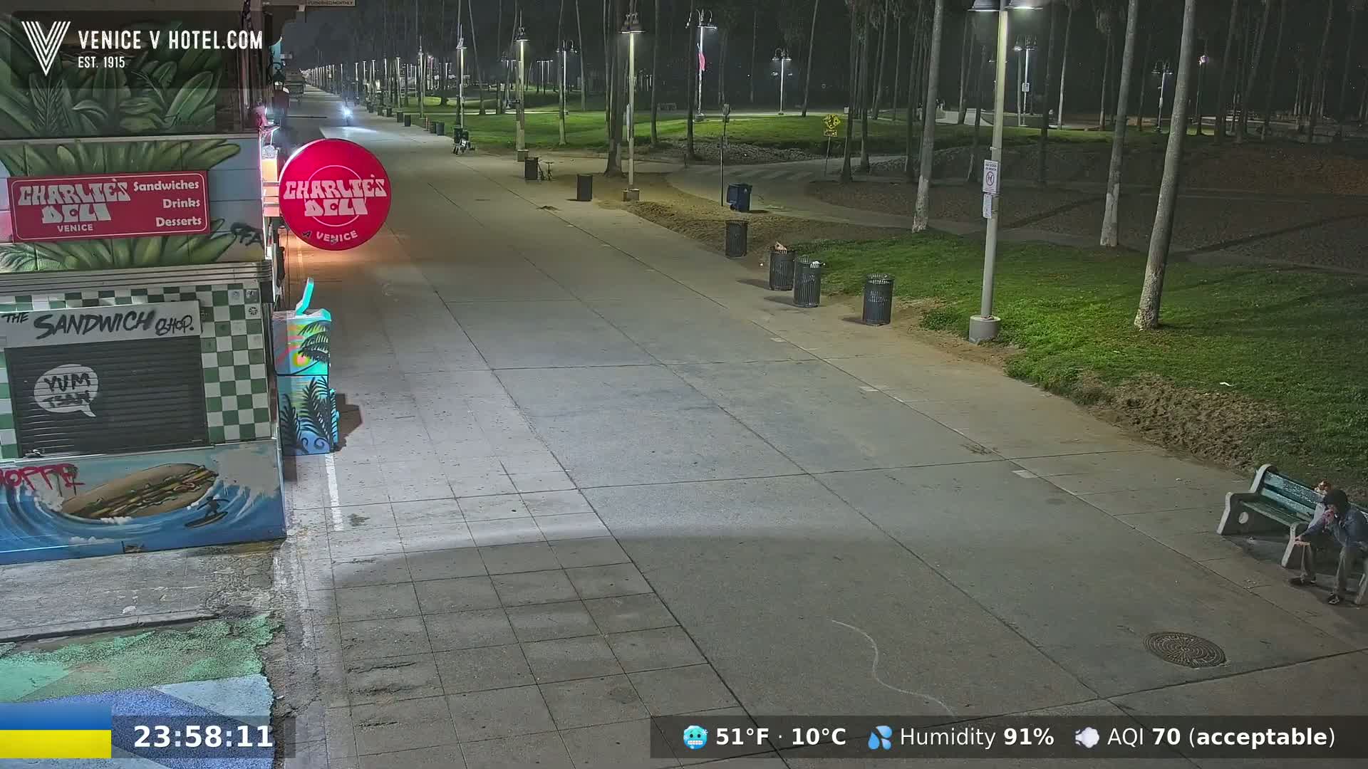 A mostly empty paved walkway at night, lined with palm trees and parkland, features a sandwich shop and a person sitting on a bench under artificial light.