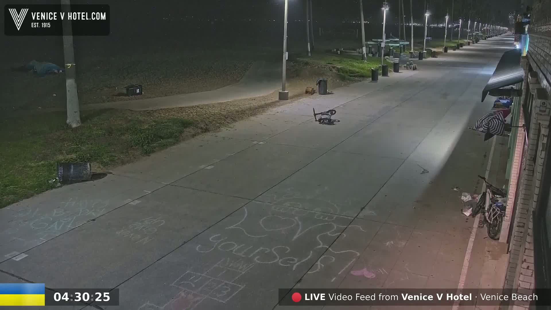 A nighttime view of a mostly empty boardwalk shows a bicycle lying on the ground, chalk drawings on the pavement, and a few scattered items of trash near a building.