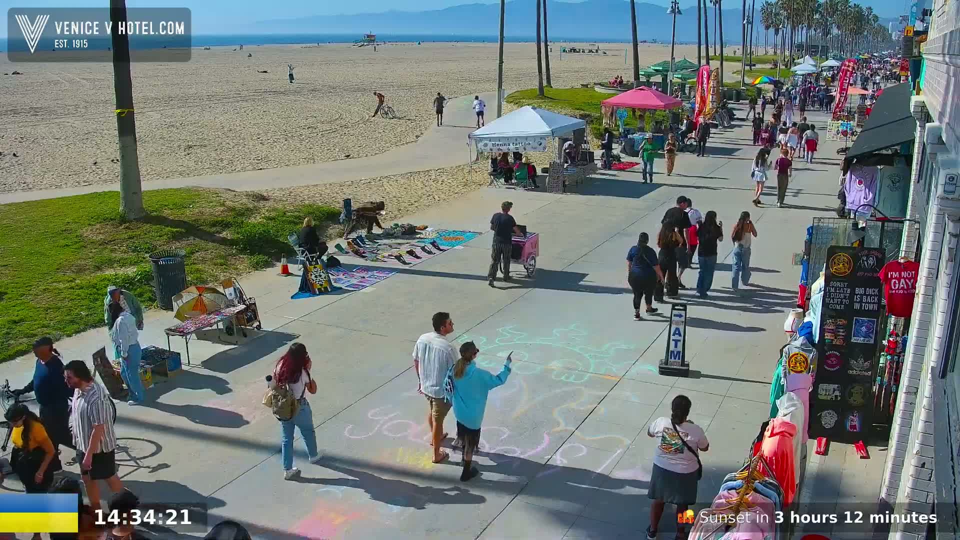 A sunny beach boardwalk scene shows people walking, vendors selling goods, and chalk drawings on the pavement.