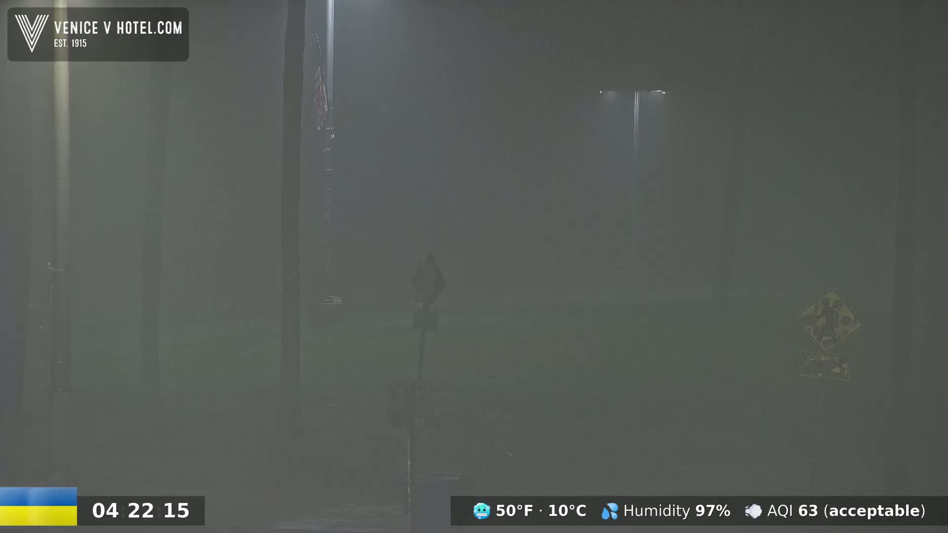 A hazy, nighttime scene shows a person standing near a street sign and light post in heavy fog or mist.