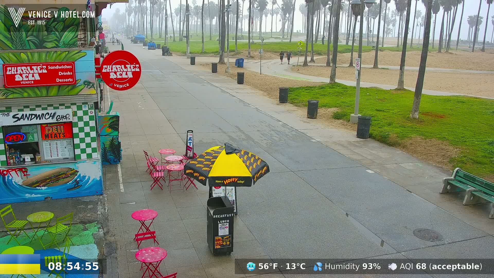 A mostly empty boardwalk on a foggy day with palm trees, outdoor seating, and a deli, with a few people visible in the distance.