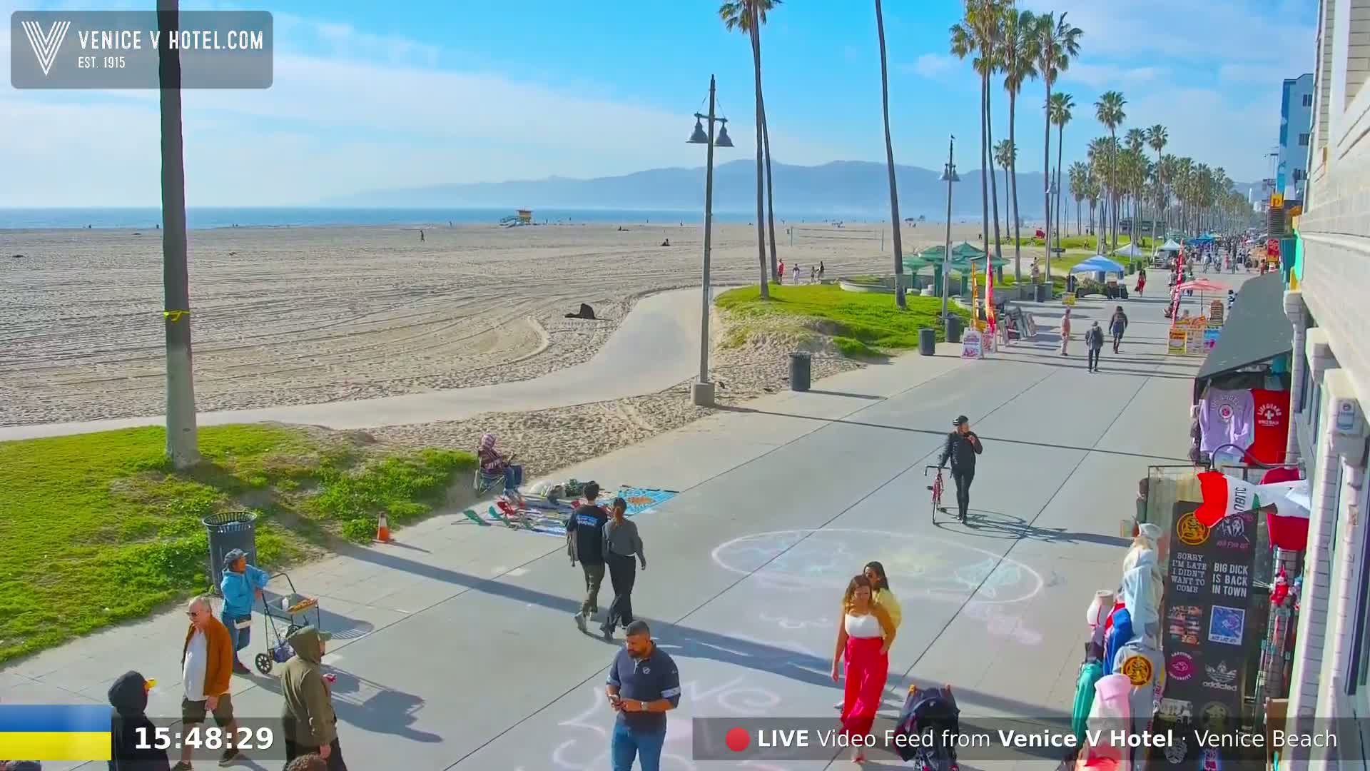 A sunny beach scene shows a wide sandy beach, a boardwalk with pedestrians and vendors, and palm trees lining the shore under a clear blue sky.