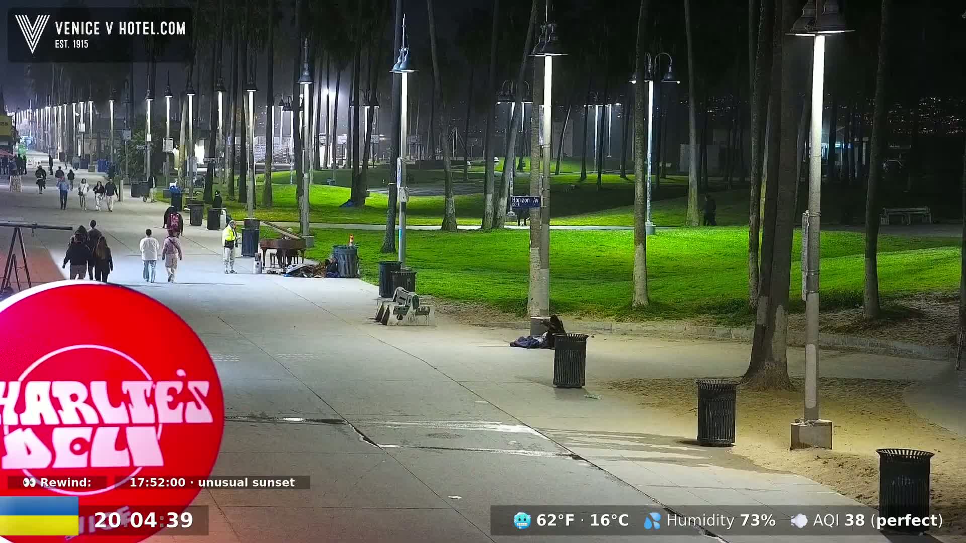A nighttime scene on a paved walkway shows people walking past palm trees and grassy areas, under illuminated lampposts, with trash cans present.