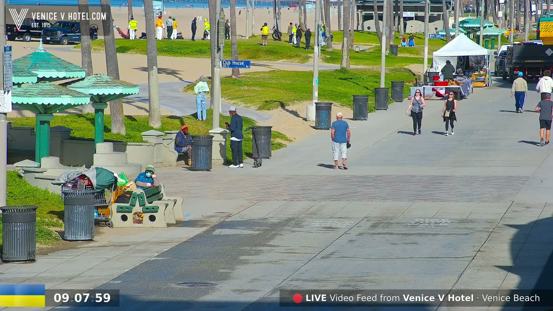 A sunny day at a beach boardwalk shows people walking, sitting, and standing near green umbrellas and trash cans, with a beach and ocean in the background.