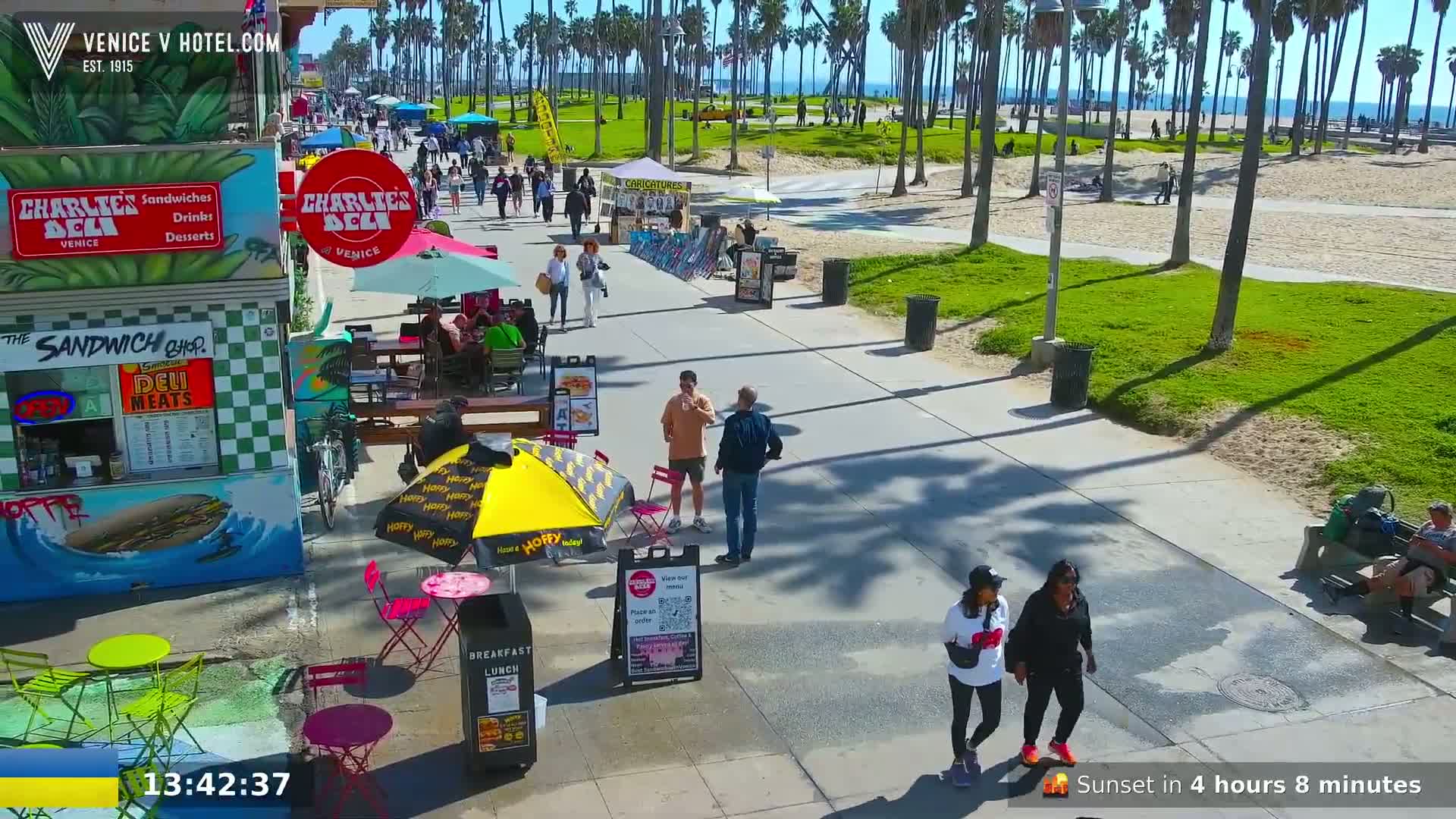 A sunny boardwalk scene with palm trees, beach views, and people walking near outdoor food vendors.