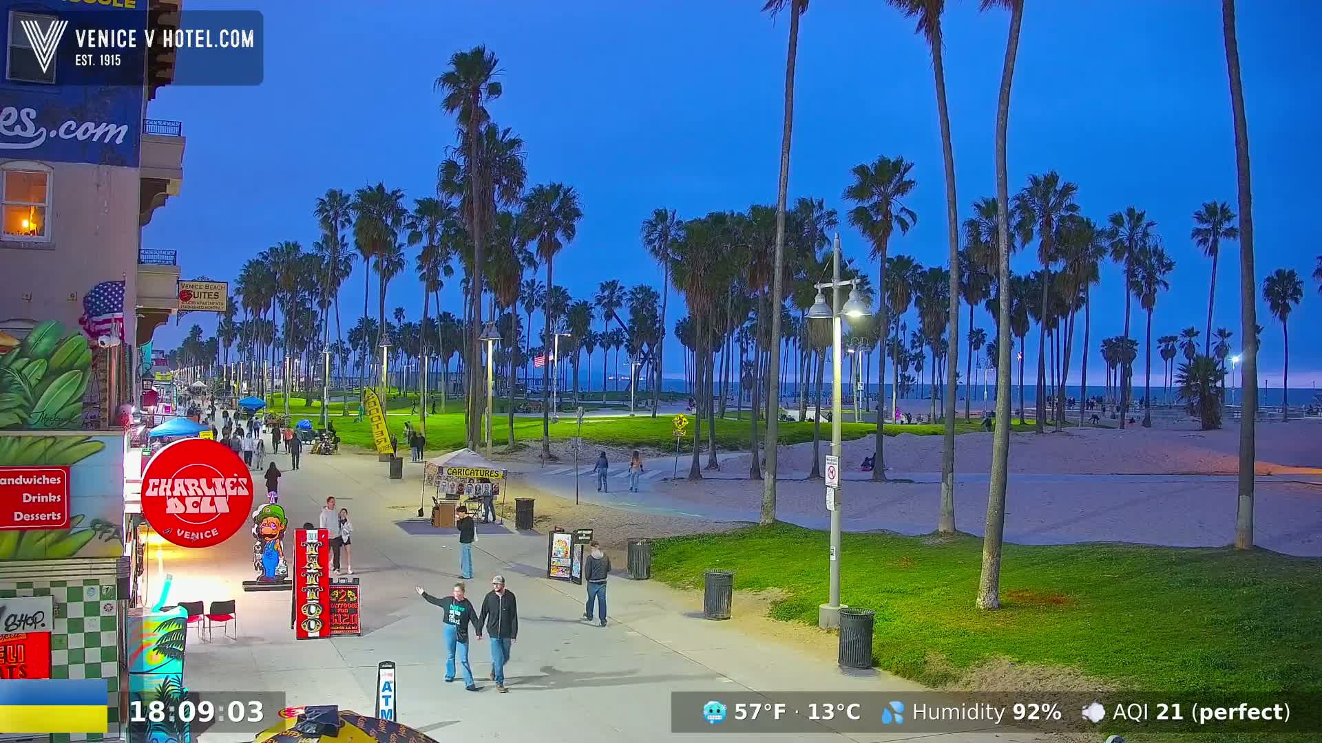 A twilight view of a beach boardwalk lined with palm trees, featuring people strolling along the path and a sandy beach in the background under a clear, dark-blue sky.