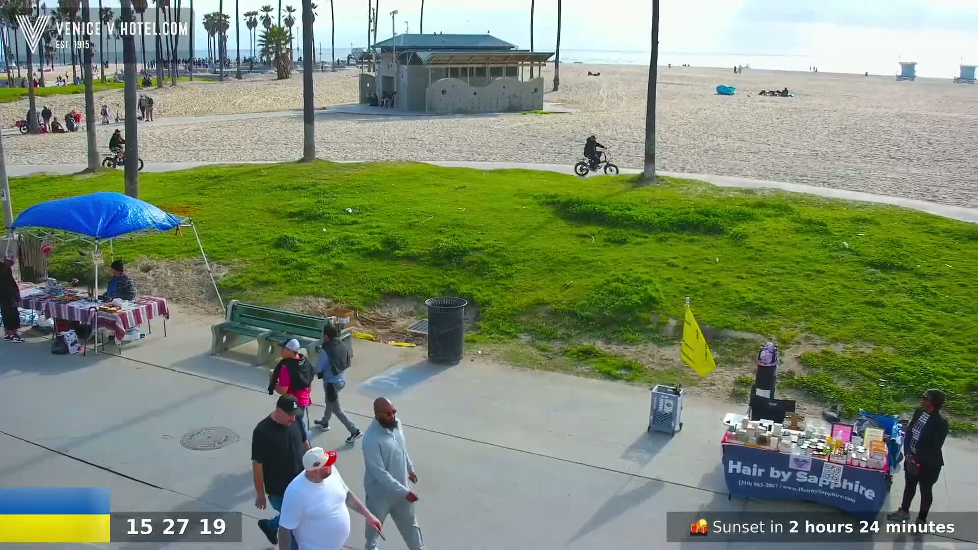 A beach scene on a sunny day shows people walking along a paved path near vendors and a grassy area, with a sandy beach and ocean in the background.