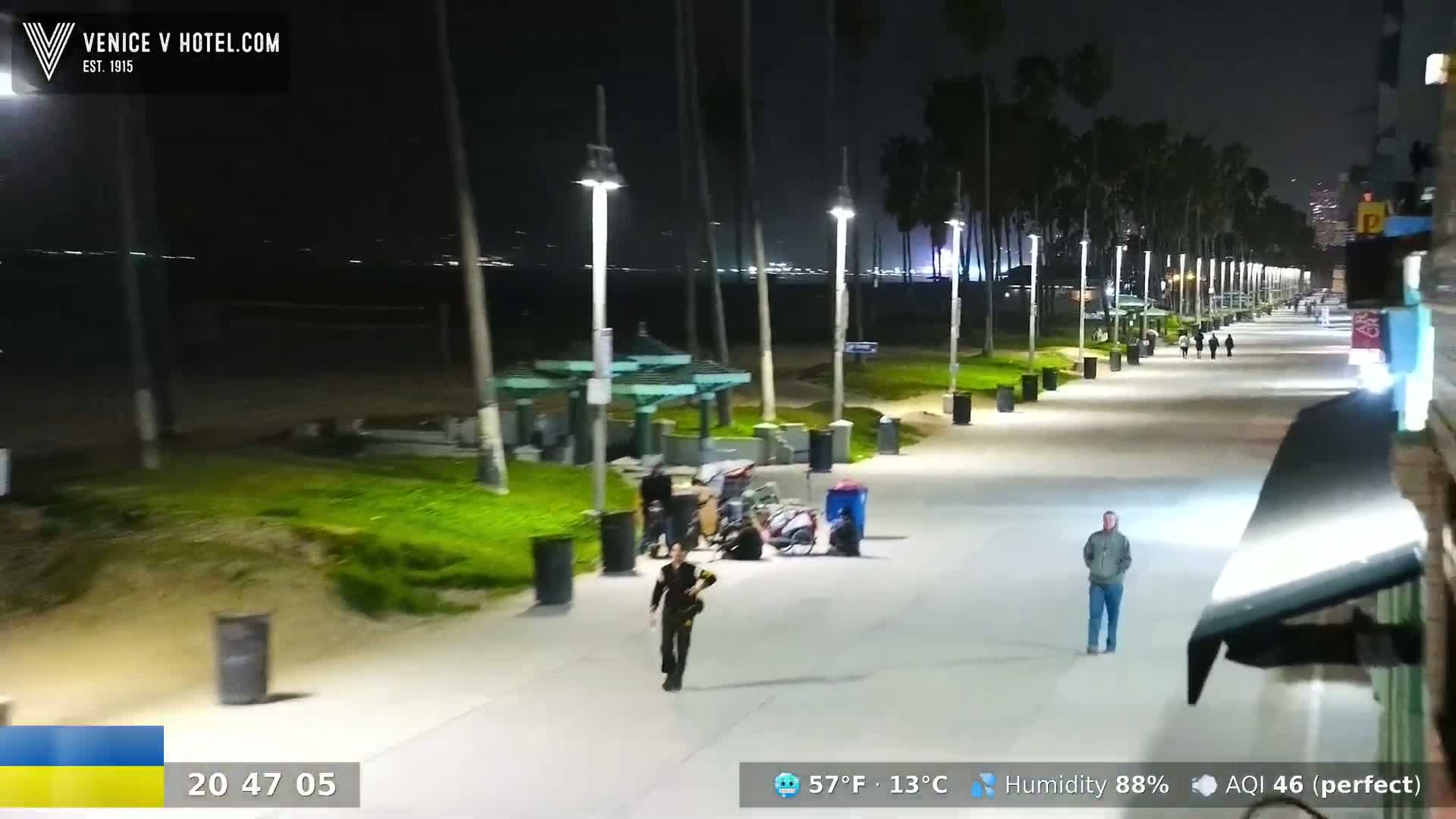 A nighttime view of a beachside boardwalk with people walking, some sitting near a gazebo, under a clear sky with palm trees lining the path.