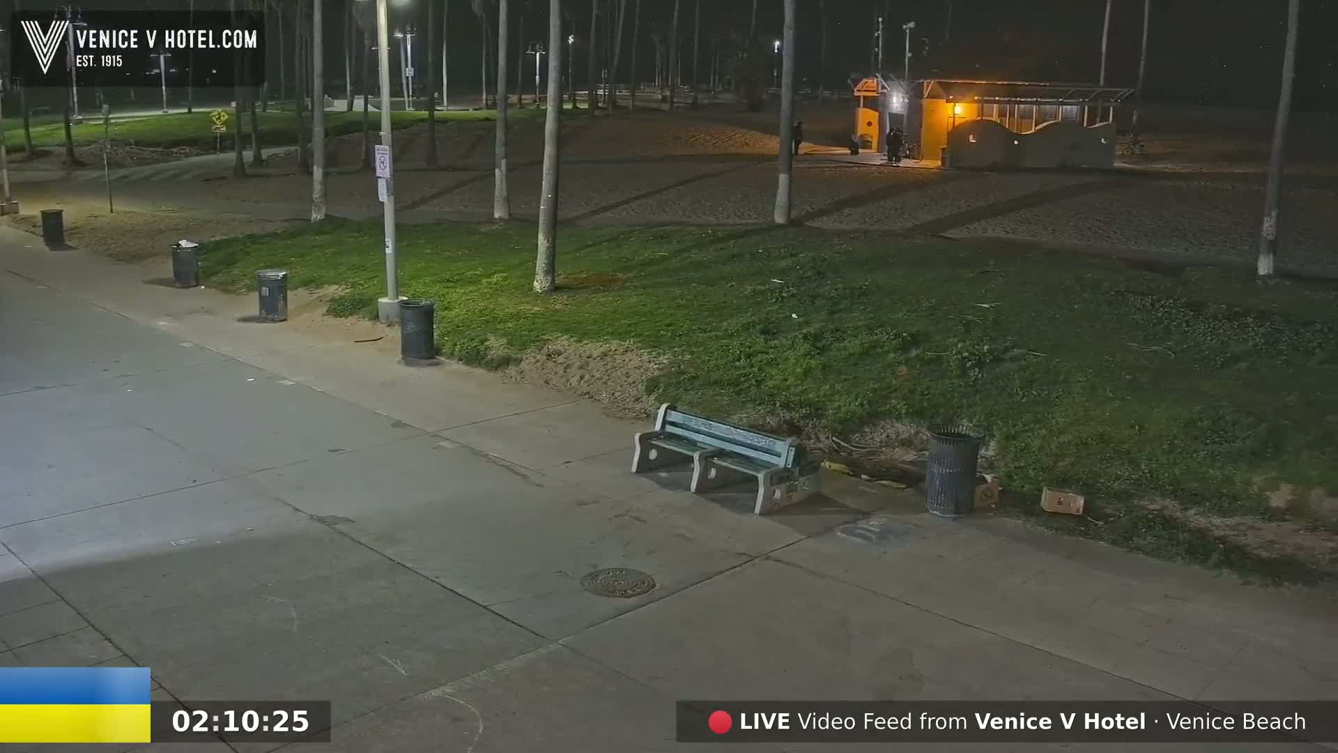 A nighttime view of a paved walkway near a beach, with grassy areas, trash cans, a park bench, and a small building in the background.