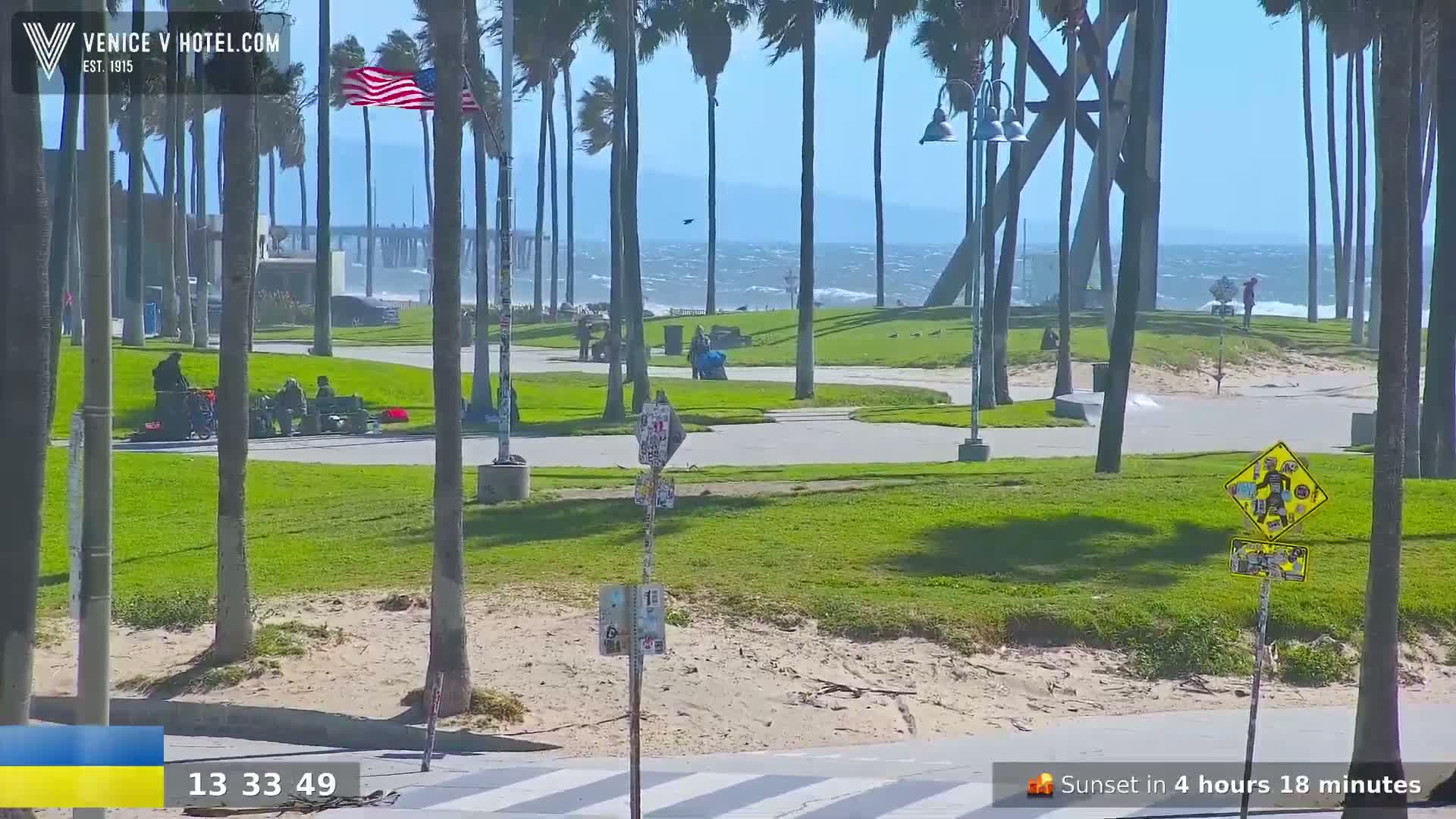 A windy beach scene shows a paved walkway, grassy areas, palm trees, and a pier in the distance; a partly cloudy, bright sunny day with a choppy ocean.