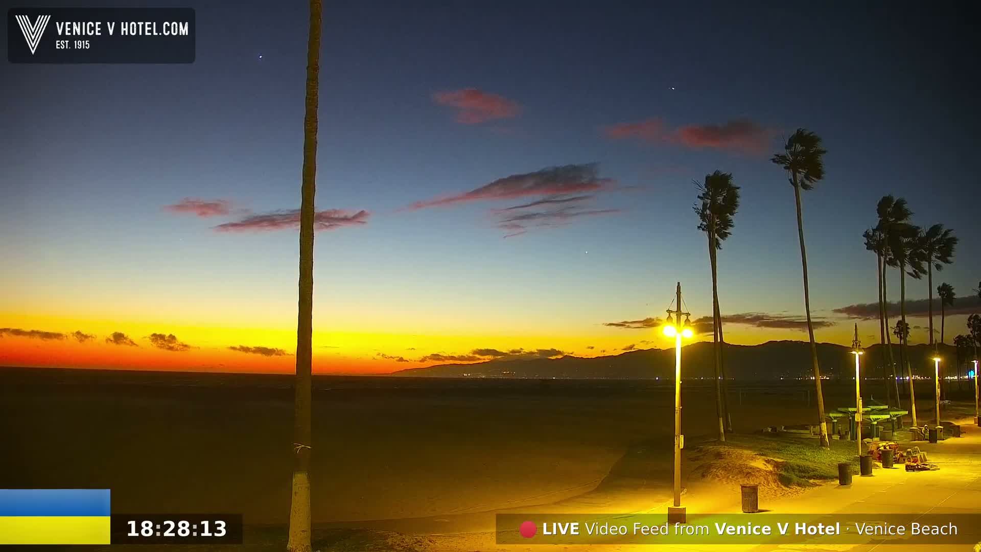 A windy sunset over a beach with palm trees, lit streetlights, and distant mountains.