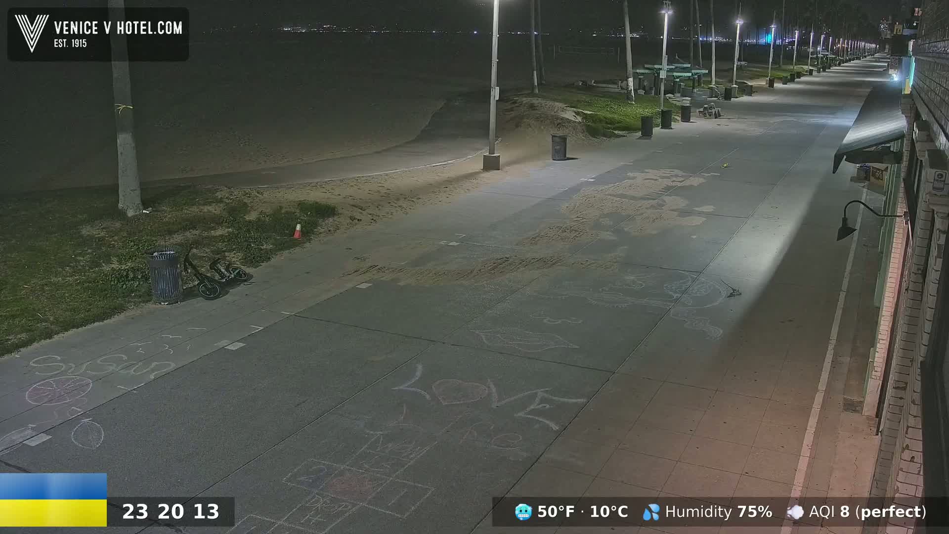 A nighttime view of a beachside boardwalk with children's chalk drawings on the pavement, a bicycle, and trash cans; the weather is clear and cool at 50°F (10°C).