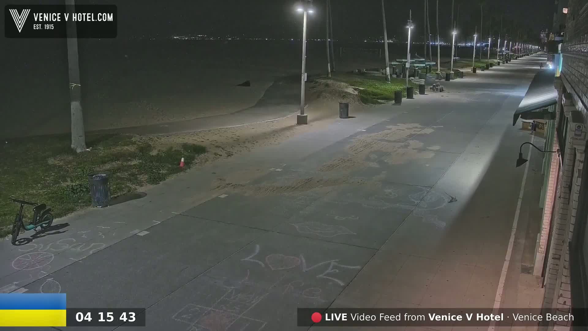 A nighttime view of a mostly empty beachfront boardwalk with children's chalk drawings on the pavement, under clear skies.