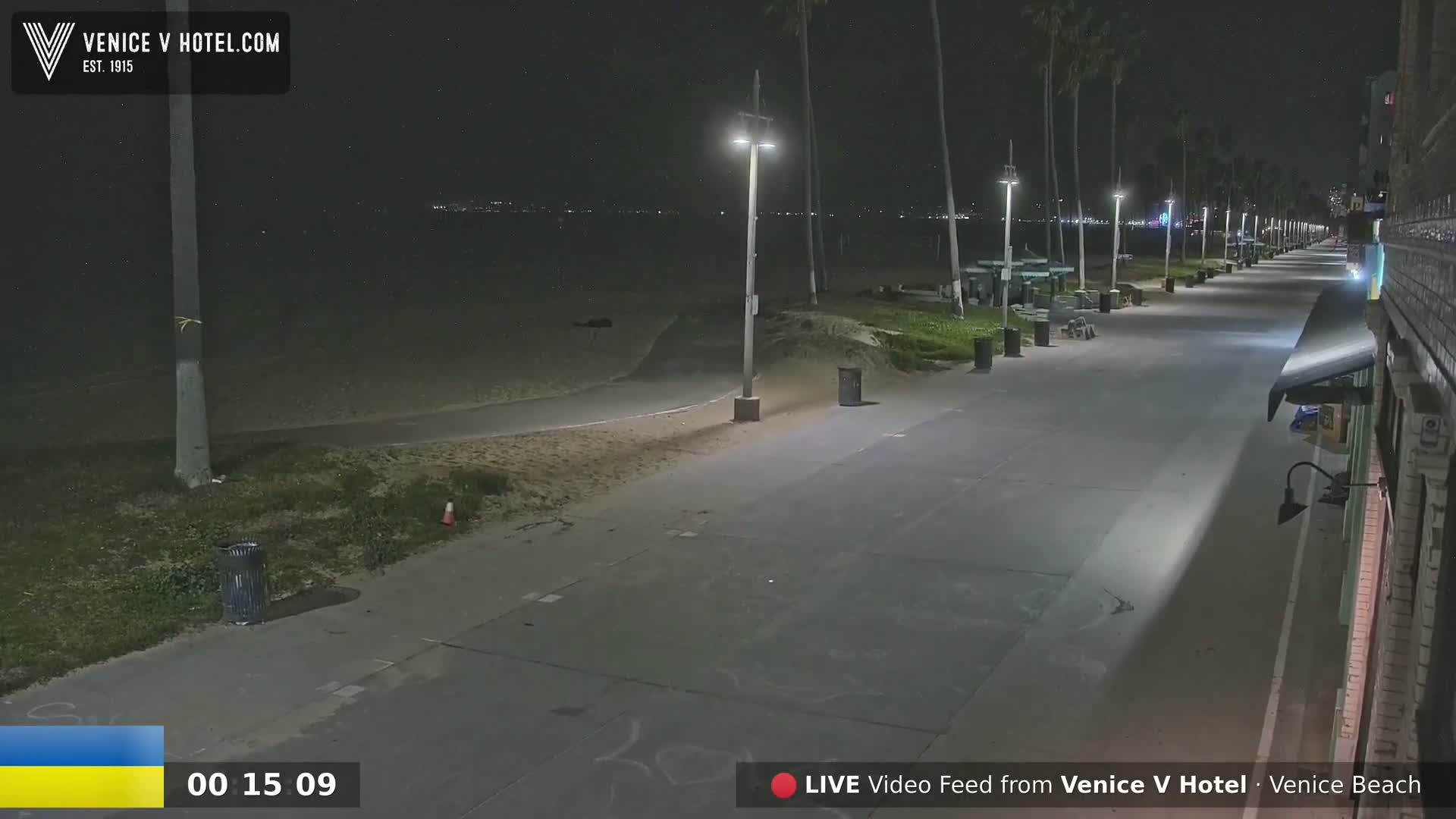 A nighttime view of a mostly empty boardwalk along a beach, illuminated by streetlights.