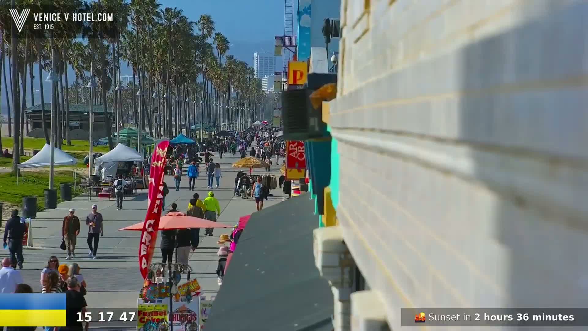 A sunny day on a boardwalk lined with palm trees shows many people walking past vendors and shops.