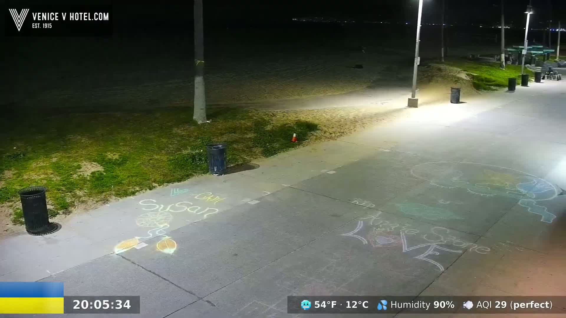 A nighttime beach boardwalk scene shows chalk drawings on the pavement, with trash cans and a grassy area nearby under brightly lit conditions and high humidity.