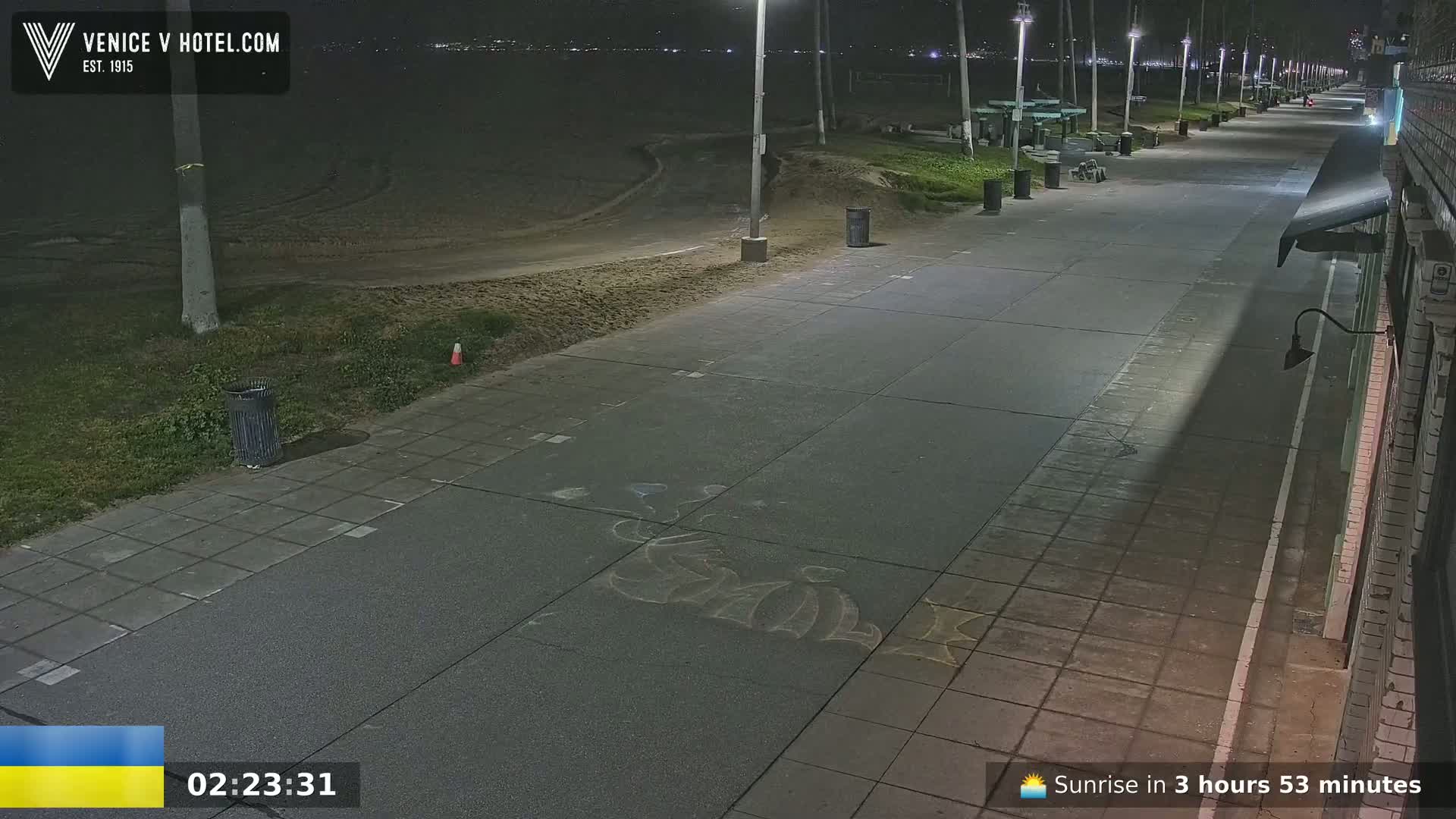 A nighttime view of a mostly empty paved walkway next to a sandy beach, lit by streetlights.