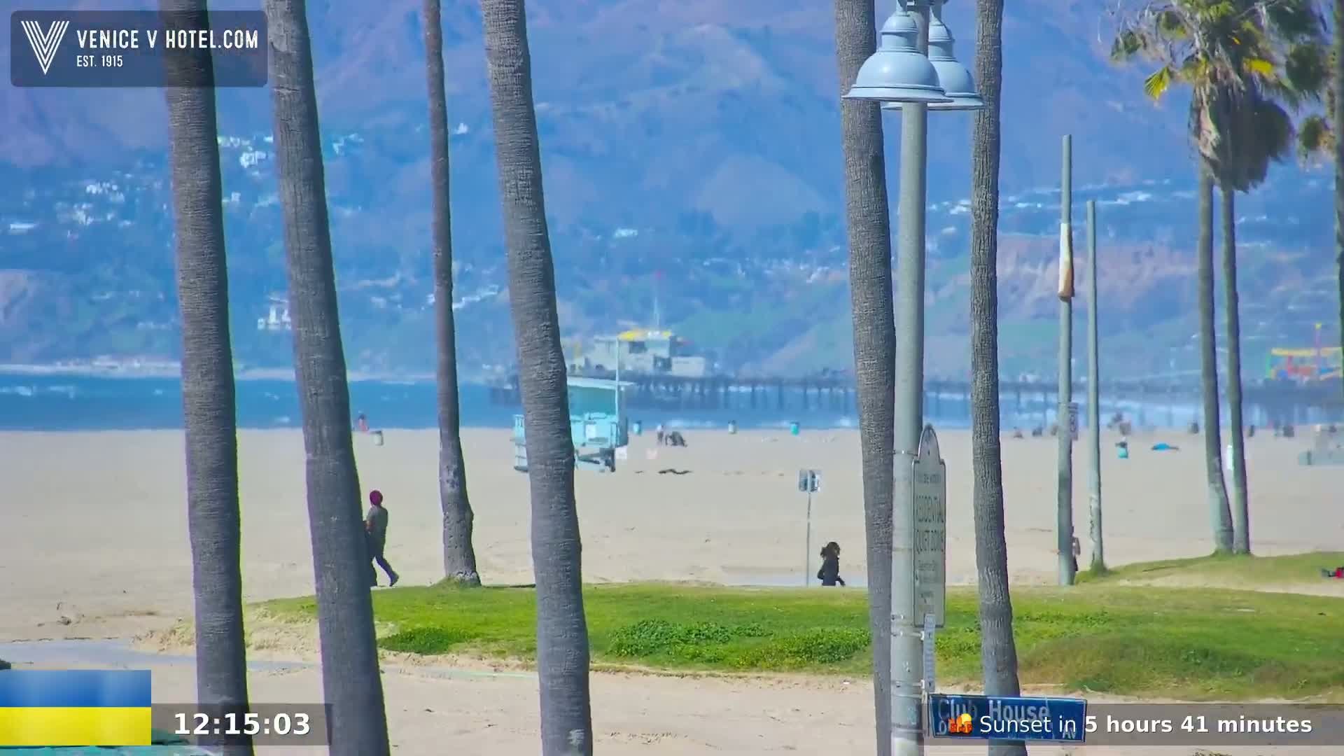 A sunny beach scene shows a sandy shore with palm trees, a pier in the distance, and a few people walking.