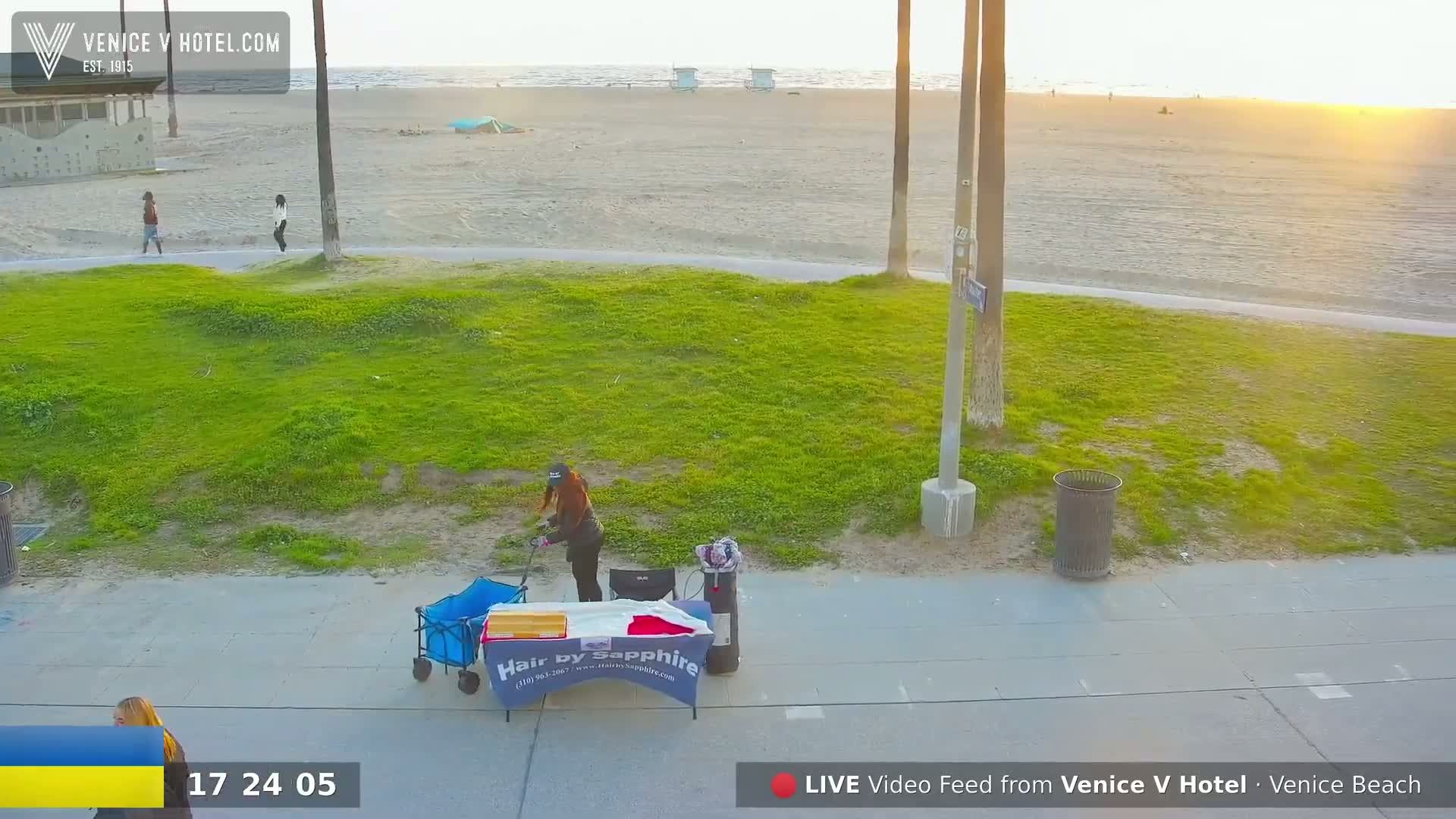 A person tends to a small table and cart on a sidewalk next to a grassy area overlooking a sandy beach at sunset.