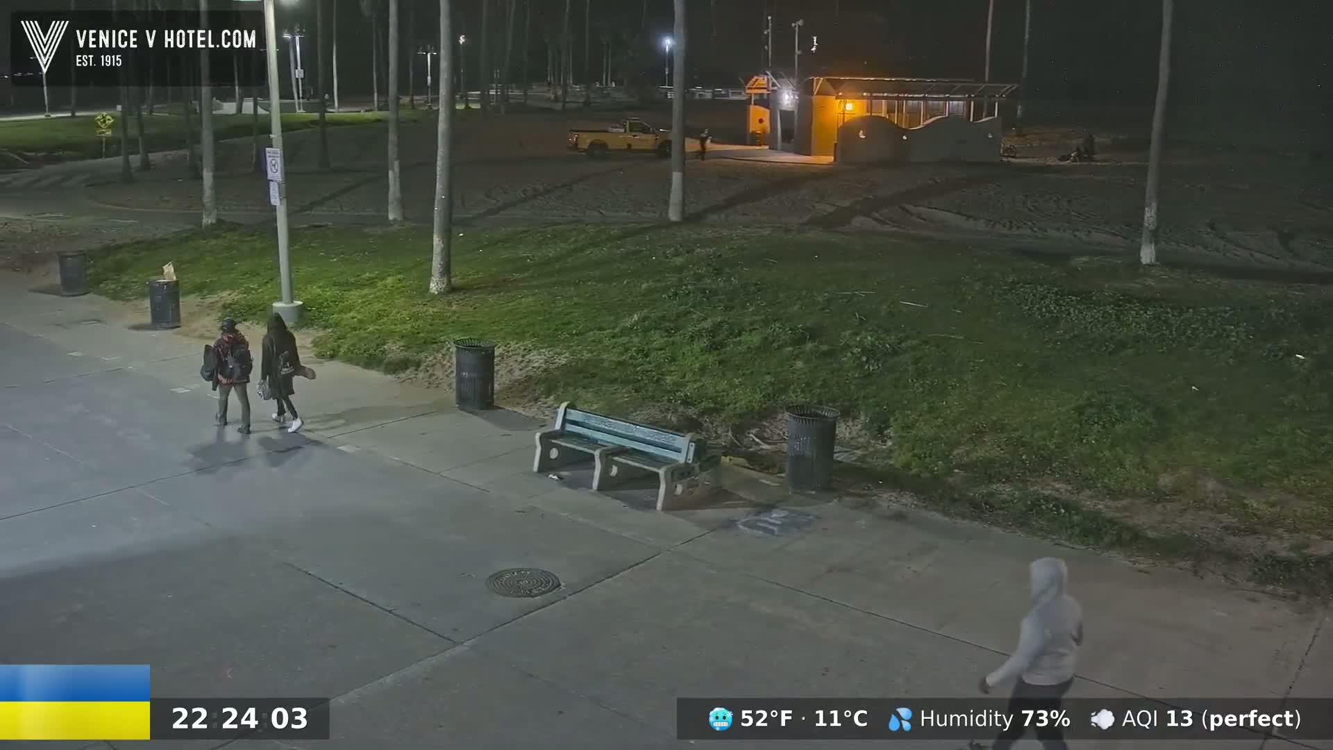At night, under a clear sky, two people walk on a paved path next to a grassy area with palm trees, a bench, and trash cans; a person walks in the distance.